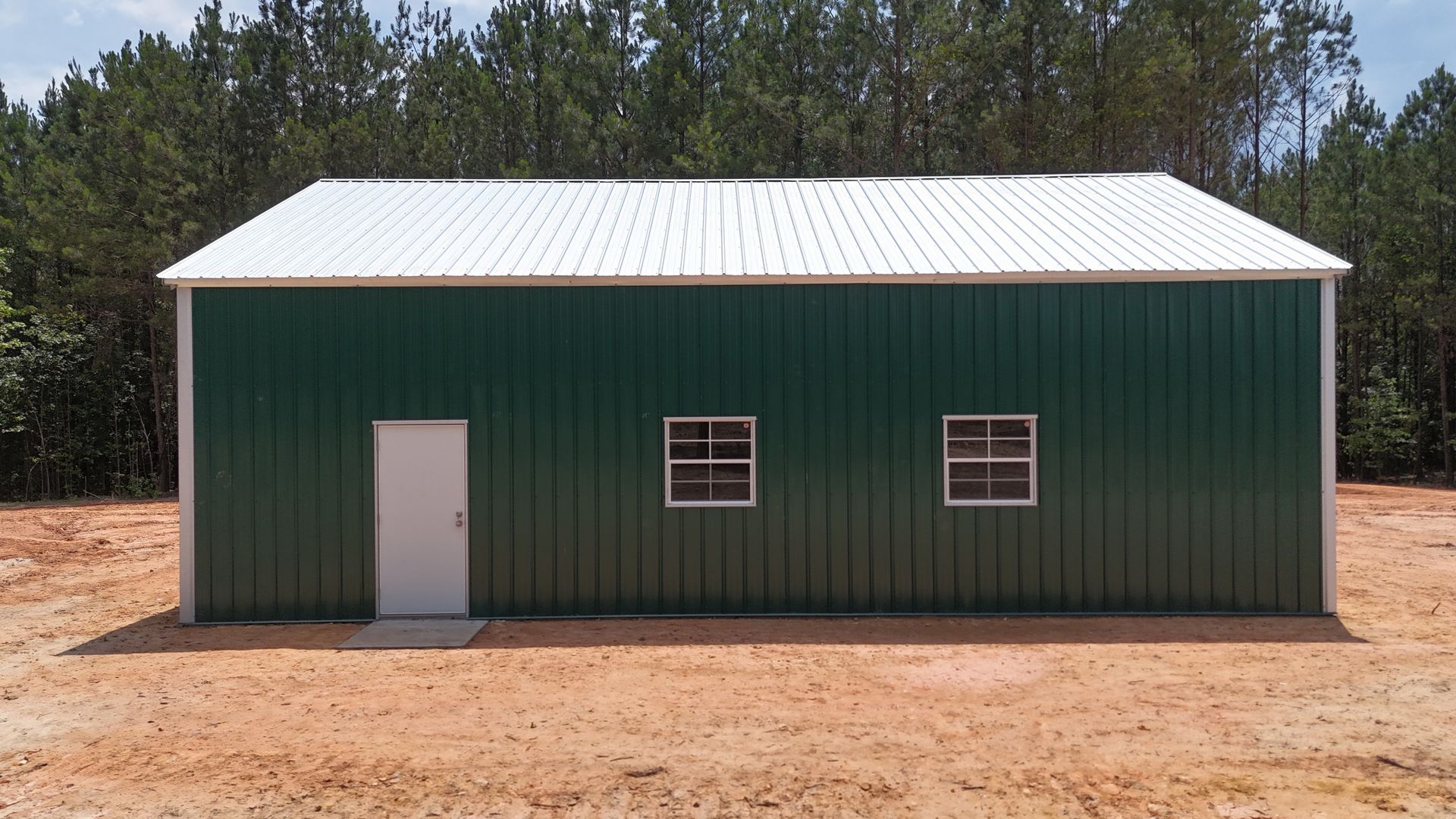 Green metal shed with white roof, white door, and two windows on a dirt lot