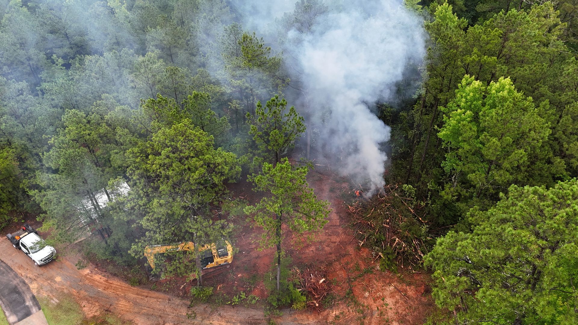 Aerial view of a forest fire sending thick white smoke over green trees and a dirt clearing.
