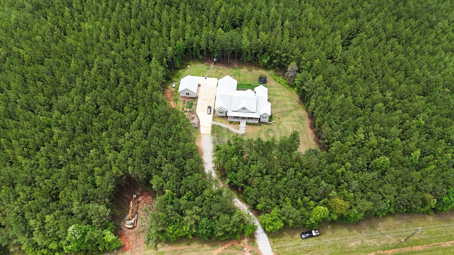 Aerial view of a white house surrounded by dense green forest, with a dirt driveway and road nearby.