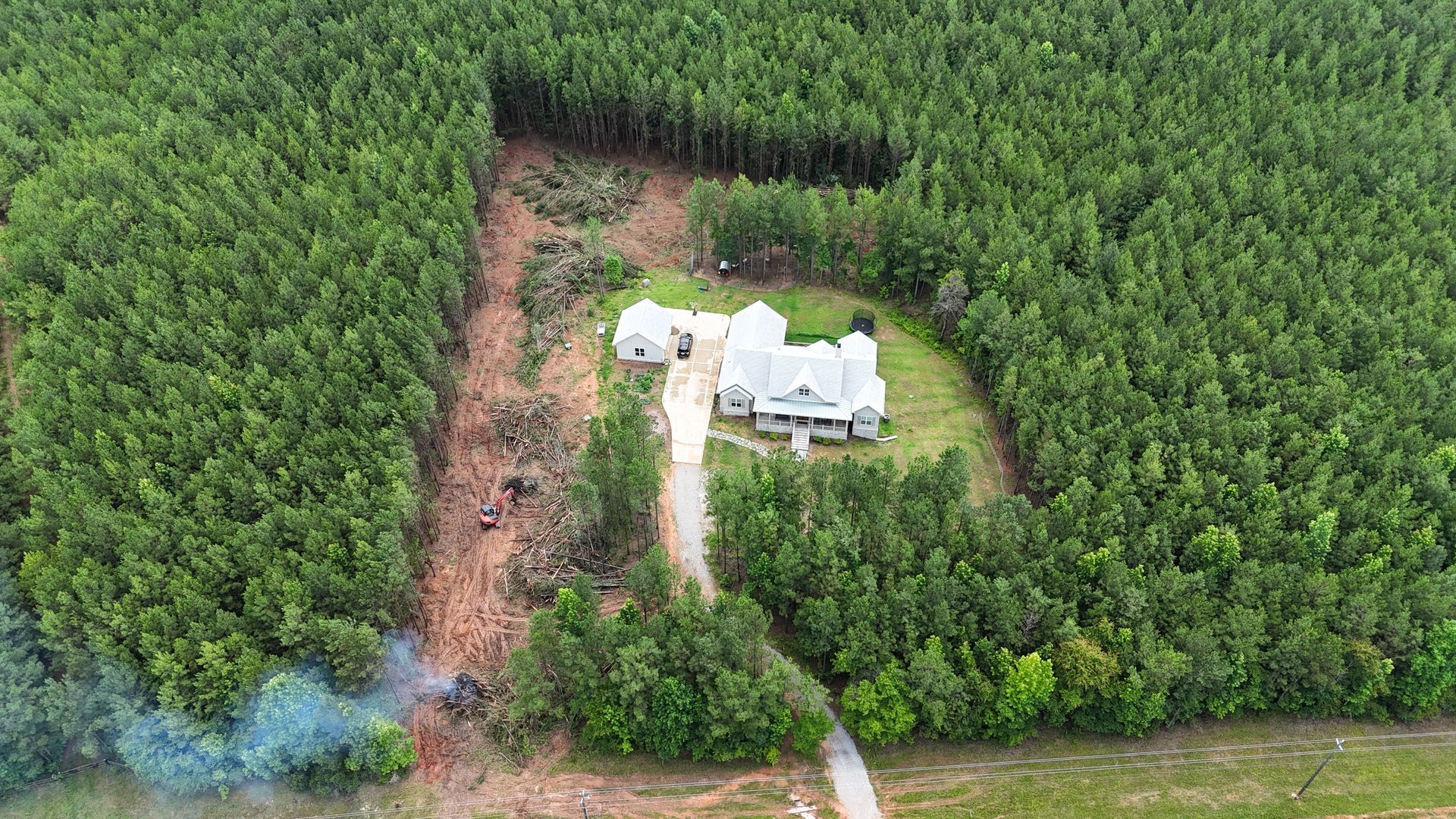 Aerial view of a white house with a dirt driveway surrounded by dense green forest.