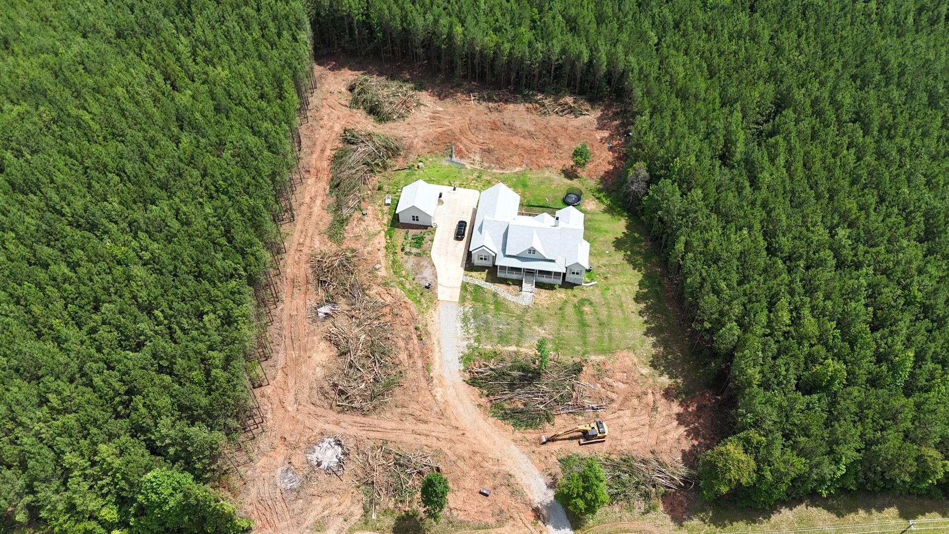 Aerial view of a white house surrounded by dense green forest with a cleared dirt area around it.