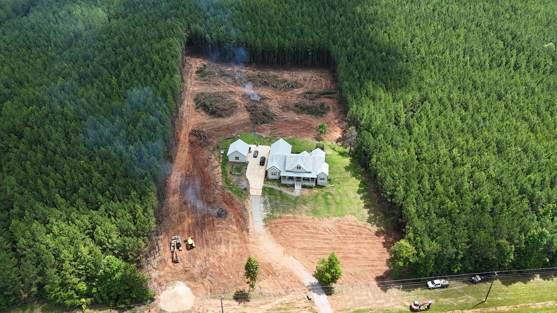 Aerial view of a white farmhouse surrounded by dense green forest and a cleared dirt area.