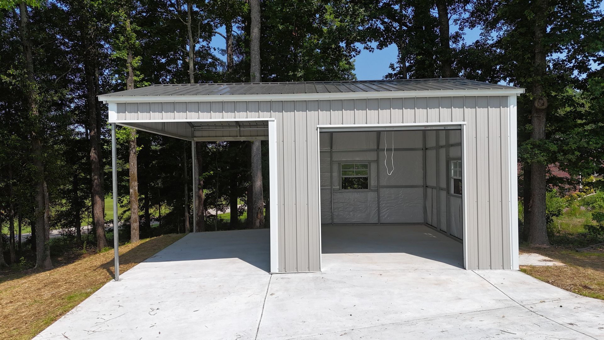 Small white metal shed with open bays and a concrete pad, set among trees in a wooded yard
