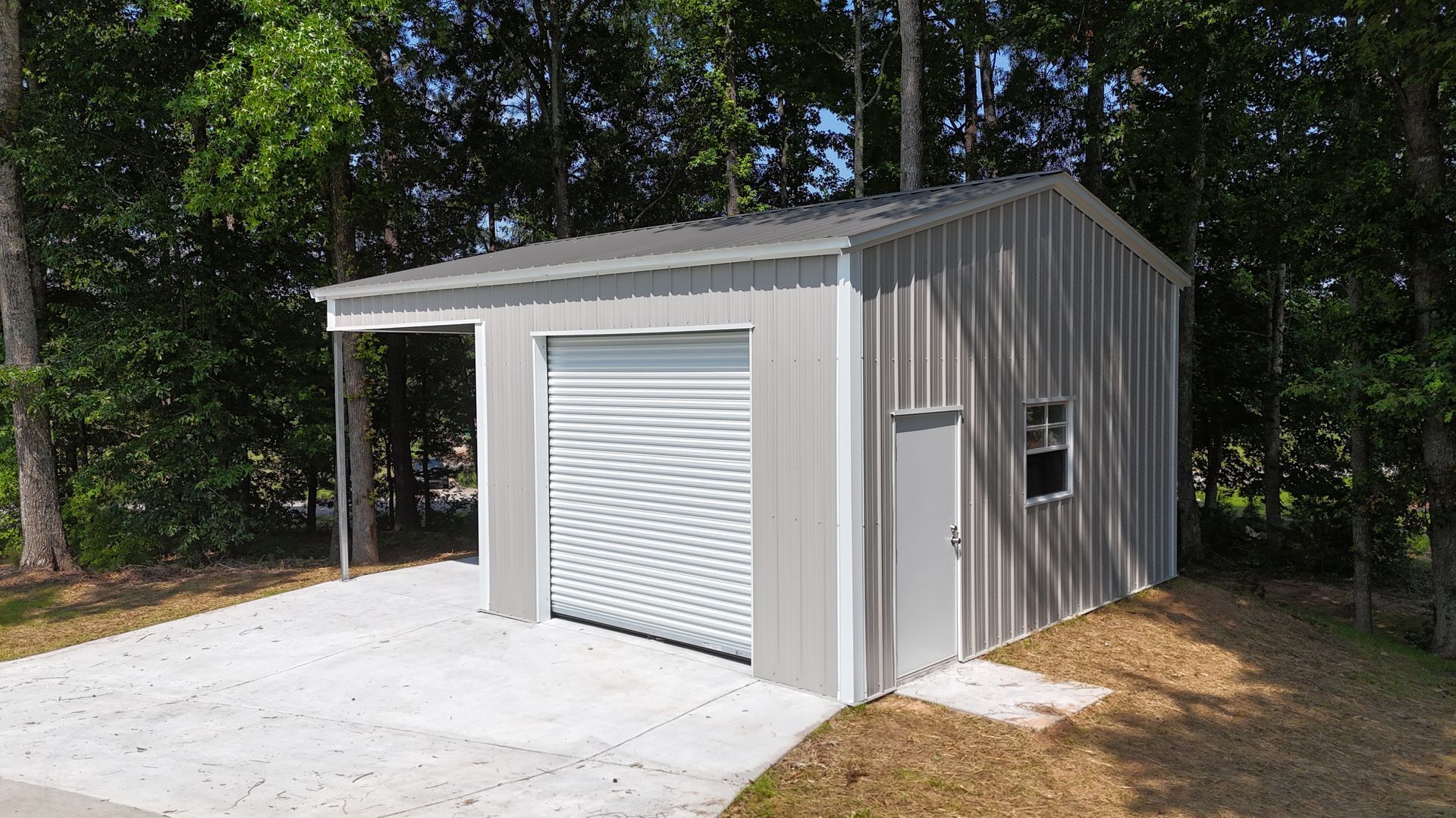 Gray metal garage/shed with roll-up door and carport on a concrete slab in a wooded area
