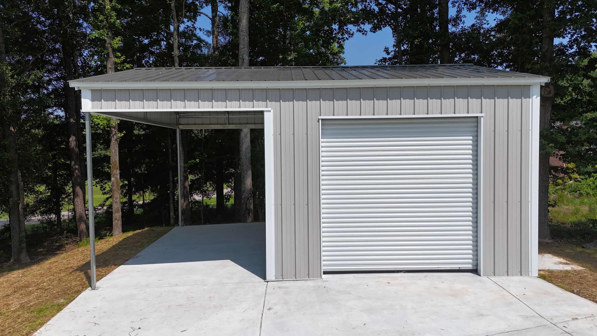 Gray metal garage with a carport and closed roll-up door on a concrete driveway, surrounded by trees.