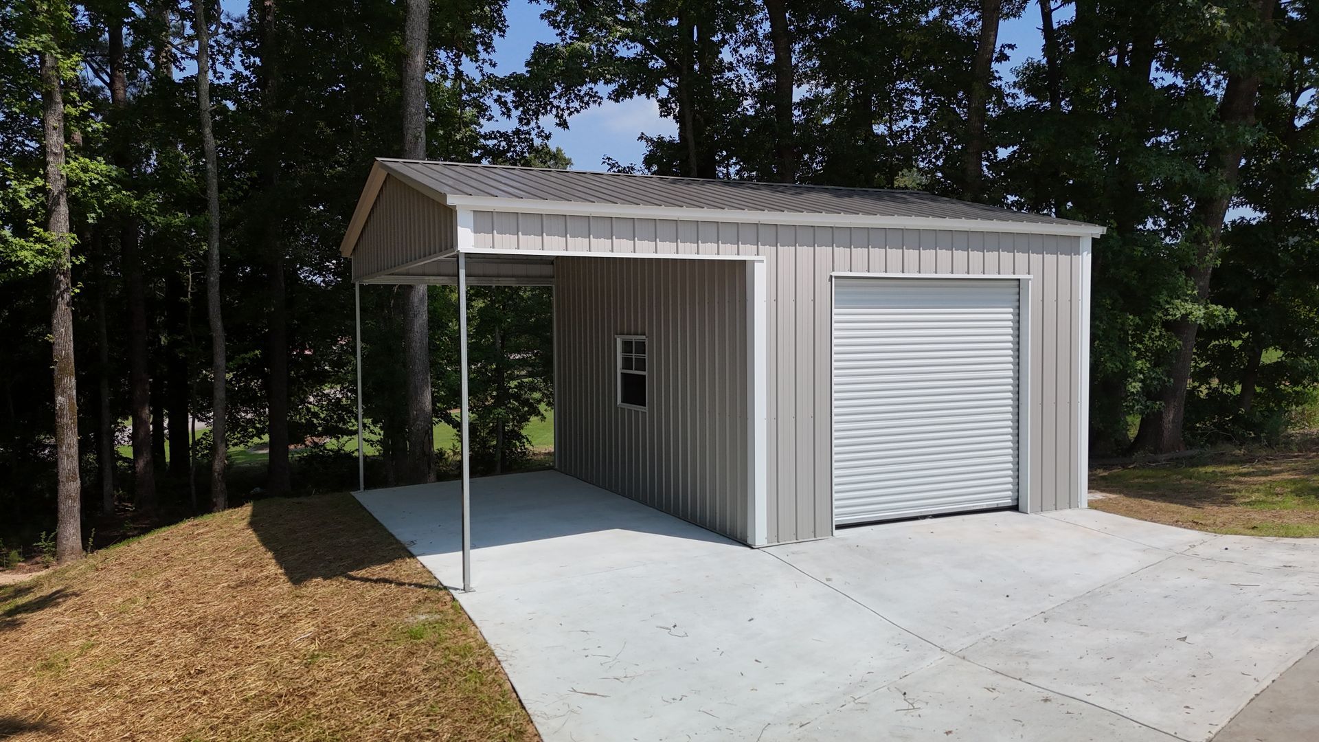 Small white metal garage or shed with a roll-up door, concrete pad, and wooded background