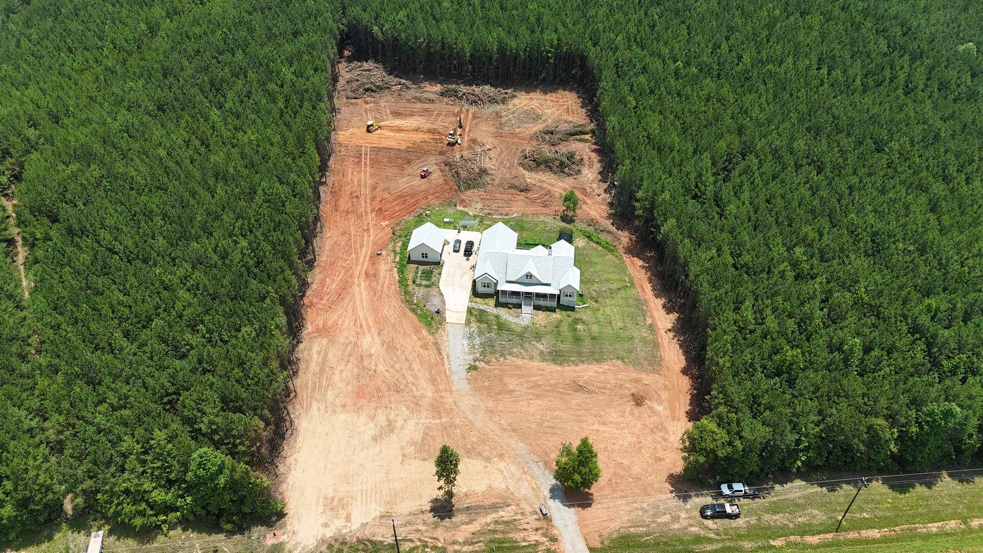A white house with a driveway sits in a cleared dirt area, surrounded by dense green forest.