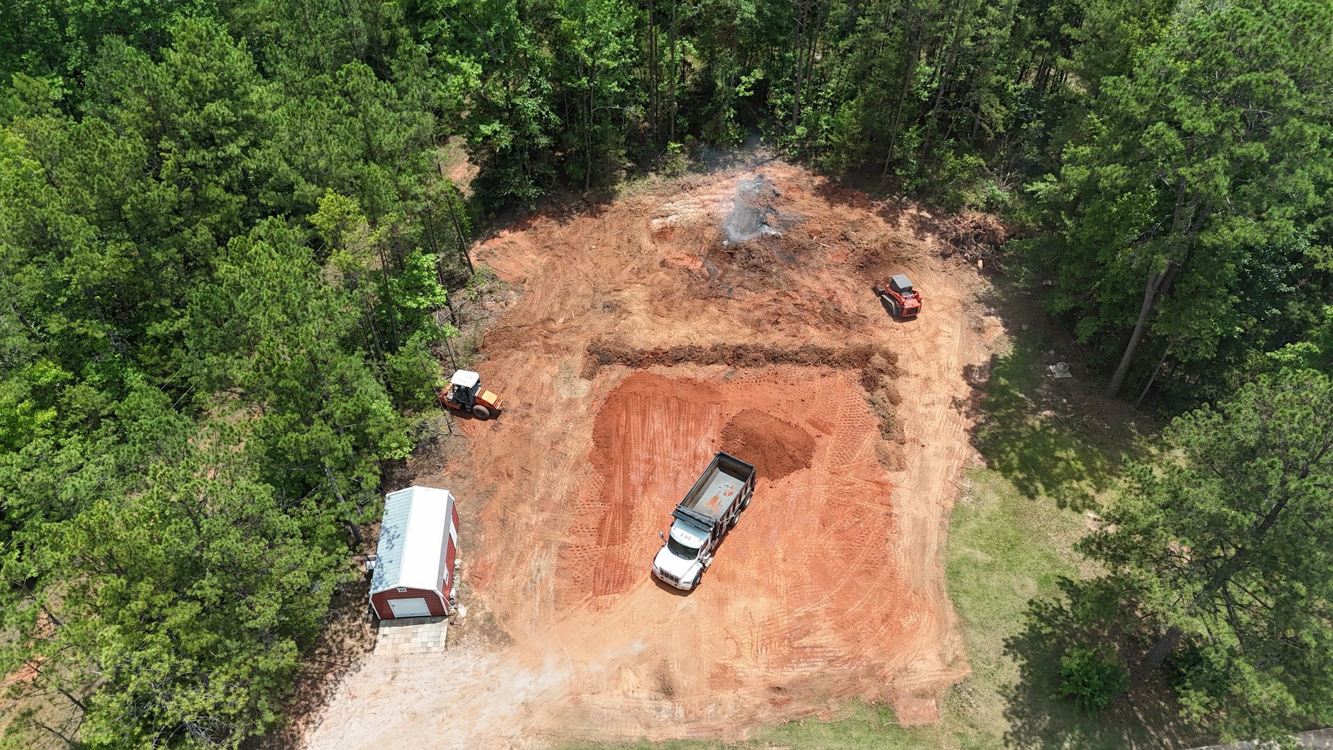 Aerial view of a dirt excavation site in a forest with two vehicles and a white trailer nearby.