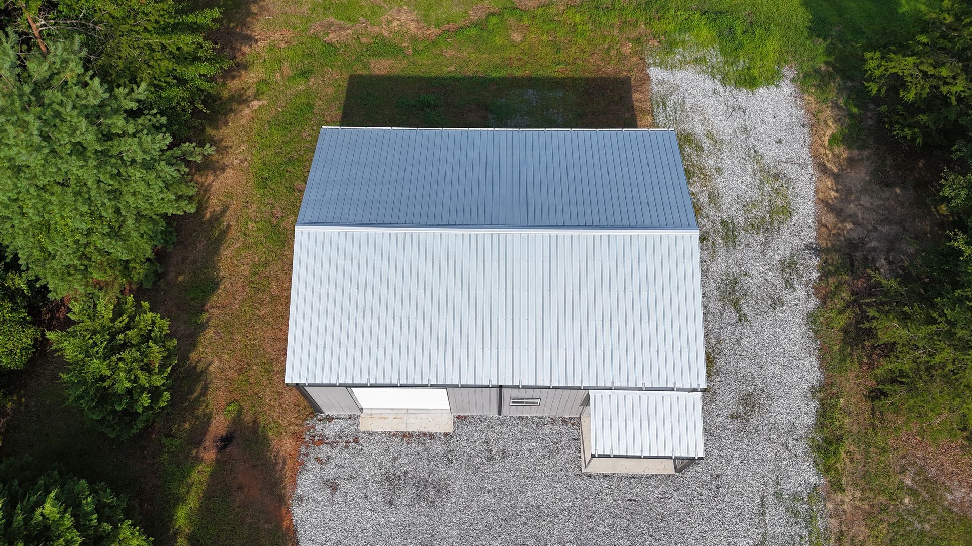 Aerial view of a small white-roofed shed beside a gravel area, surrounded by trees and greenery.