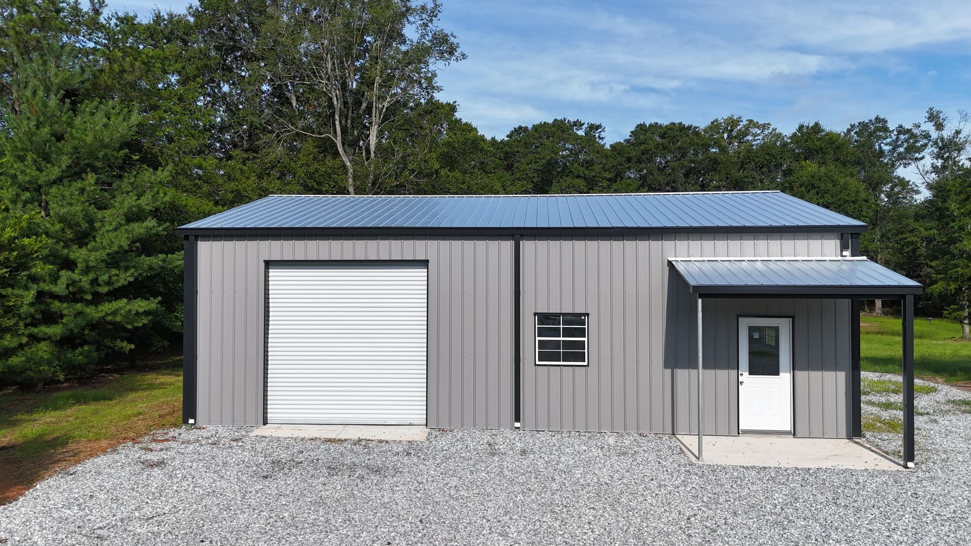 Gray metal garage building with white roll-up door and small entry porch on a gravel lot.