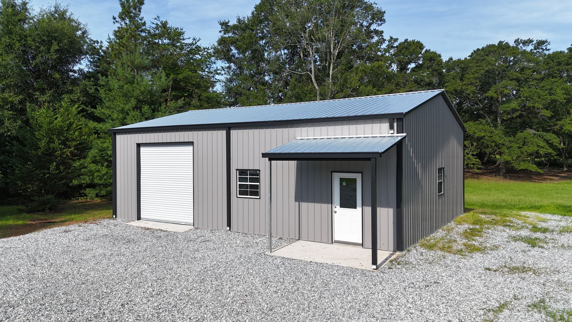 Gray metal garage building with white roll-up door and small entry door on gravel lot, surrounded by trees.