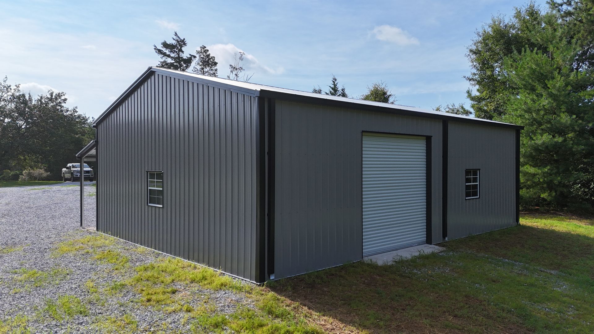 Dark gray metal storage building with a roll-up door, sitting on gravel near trees and grass.