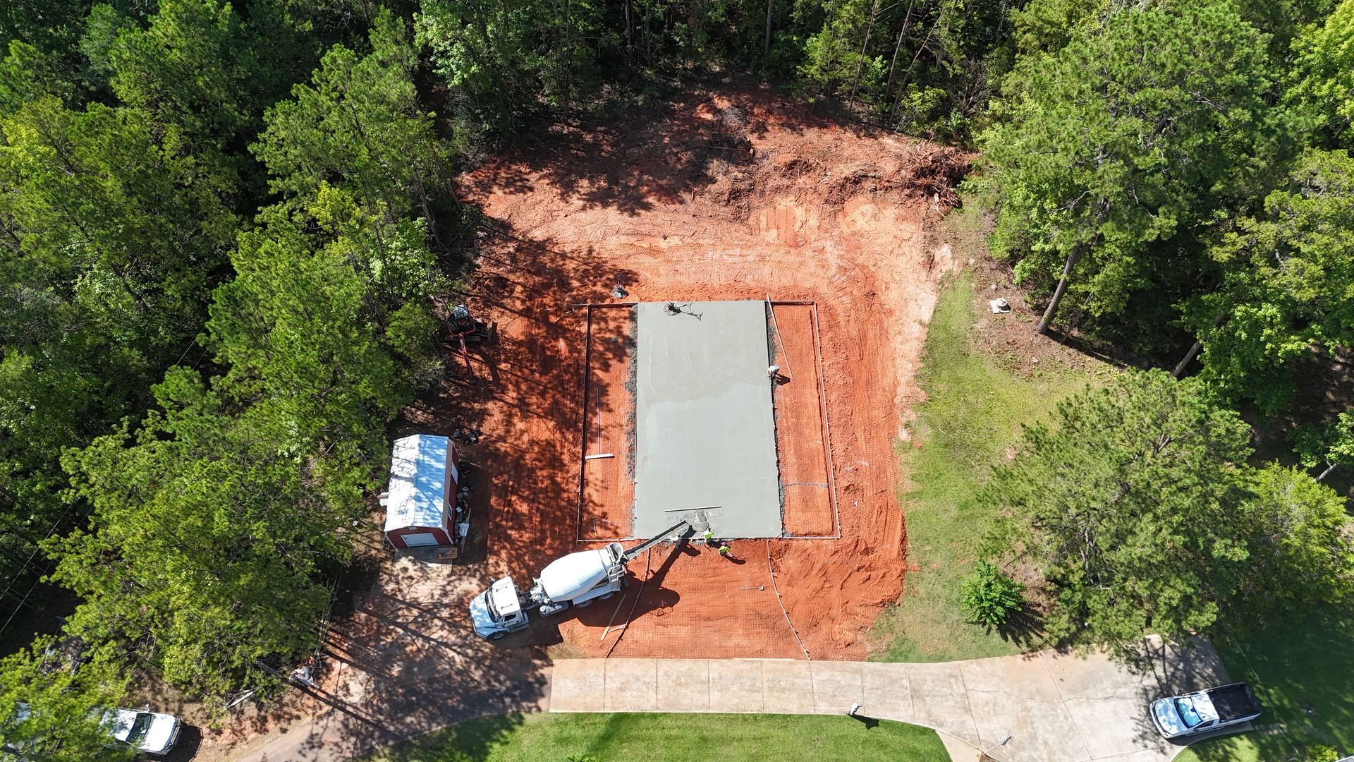 Aerial view of a small construction site with a concrete slab, red dirt, and surrounding green trees.