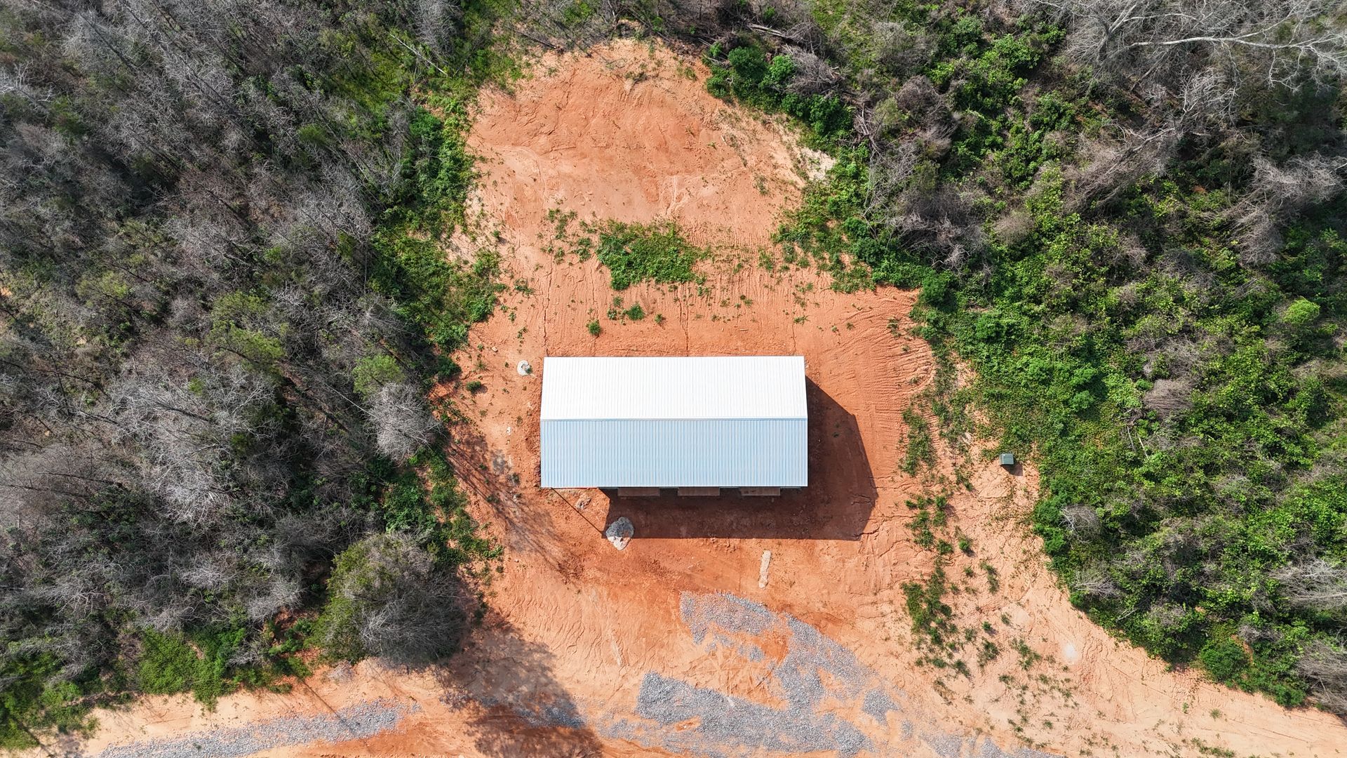 Aerial view of a small white-roofed shed on a red dirt clearing surrounded by trees and brush.
