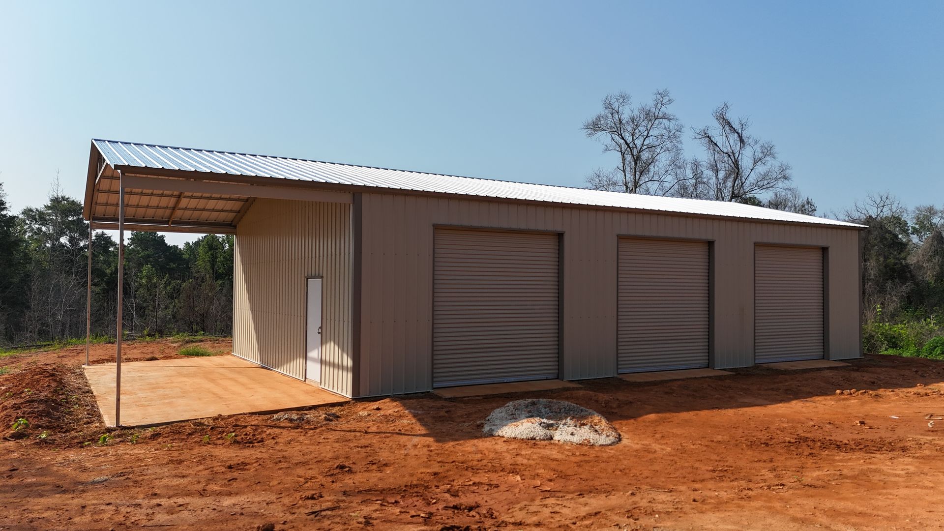 Small modern building with a slanted roof and screened windows on red dirt, beside trees under a clear sky