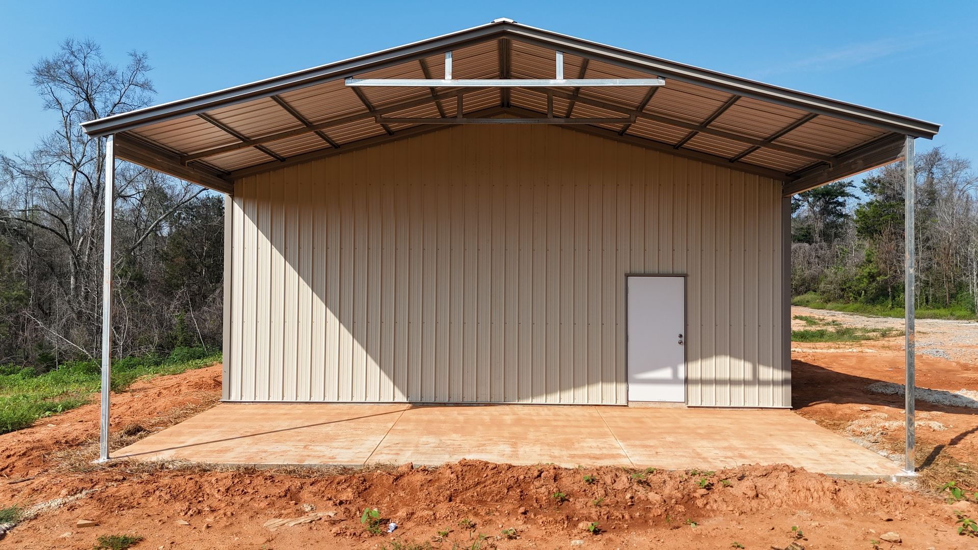 Small beige metal shed with a large roof, standing on bare red dirt in a rural setting.
