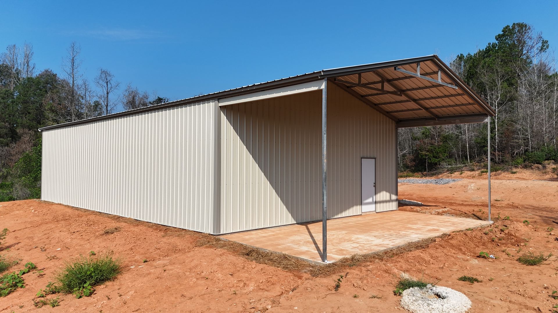 Small beige metal shed with a covered porch on a red dirt lot under a clear blue sky