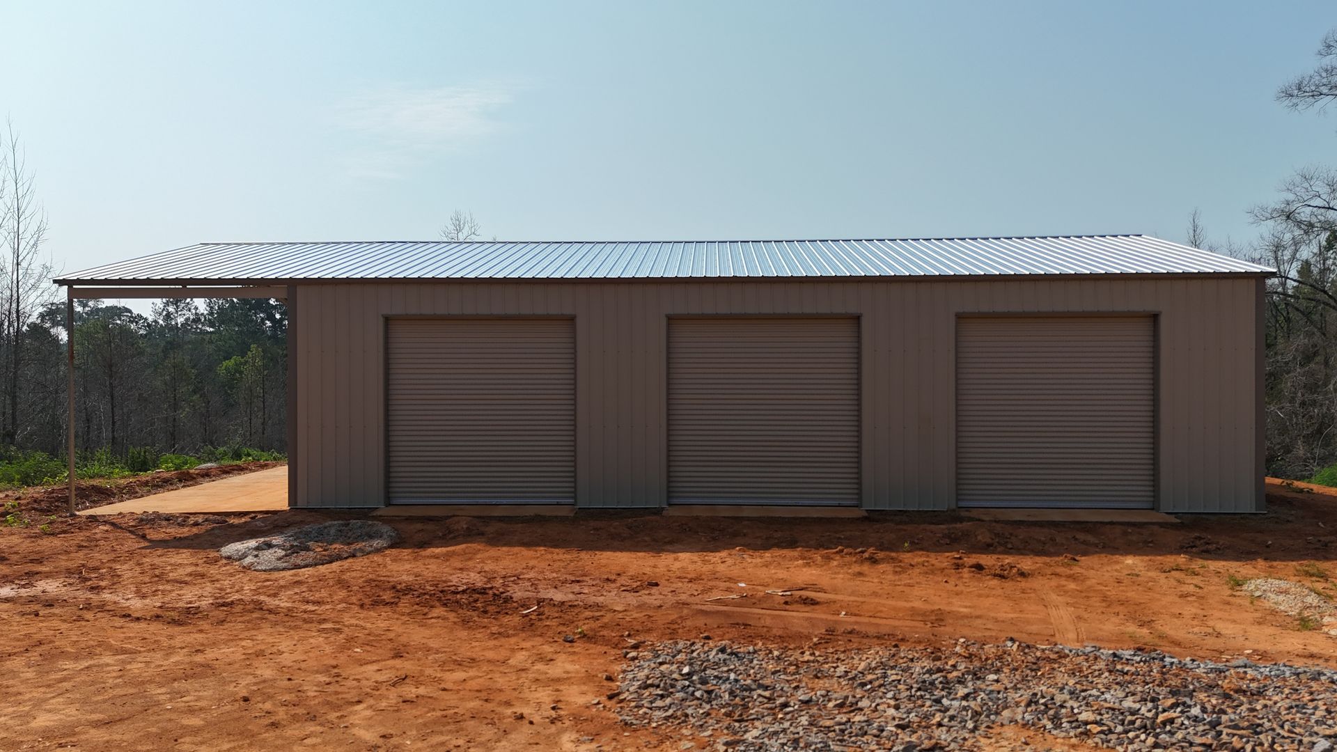 Single-story metal garage with three roll-up doors on red dirt under a clear sky
