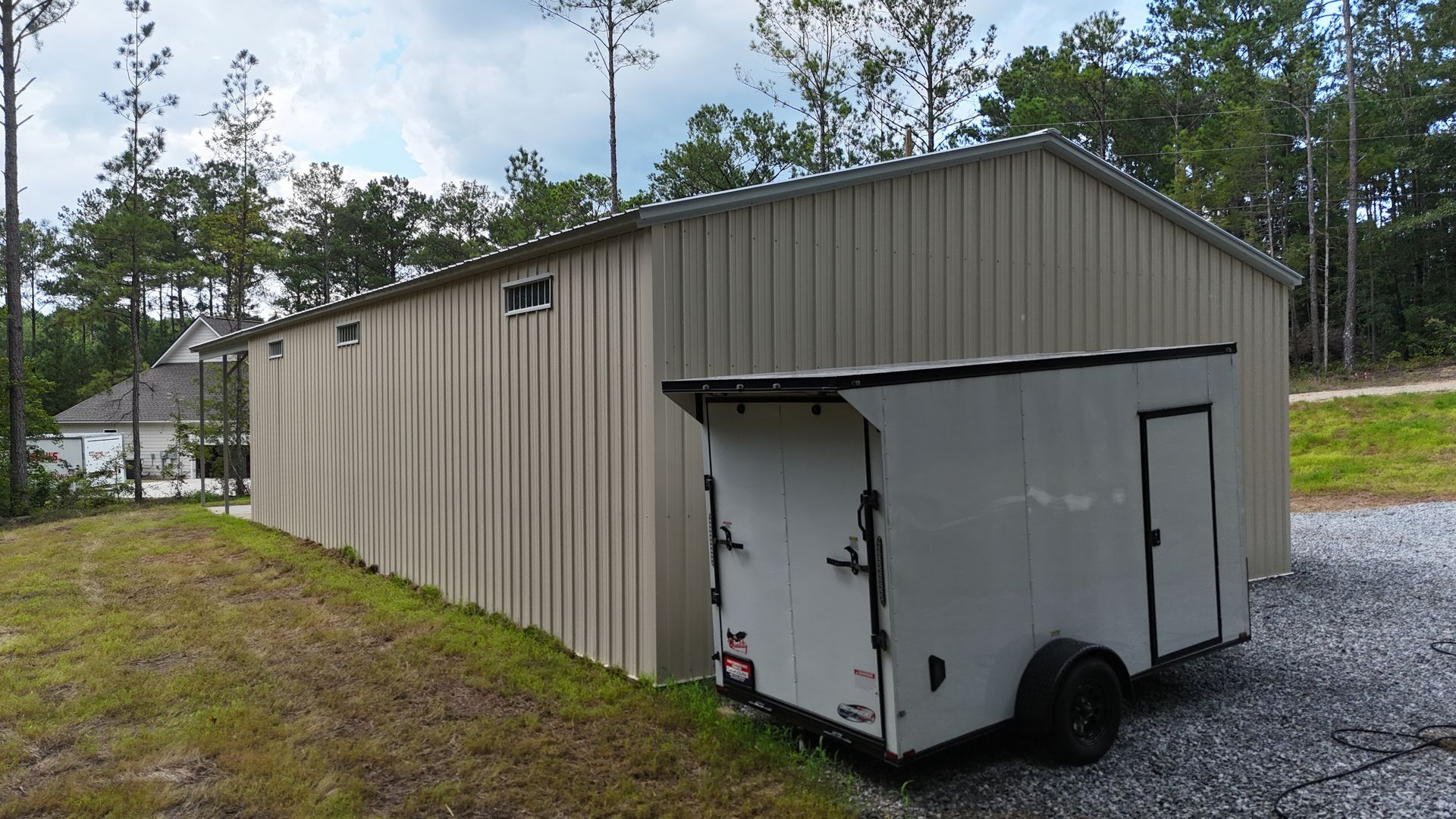 White enclosed trailer parked beside a beige metal building on a gravel drive, with trees in the background