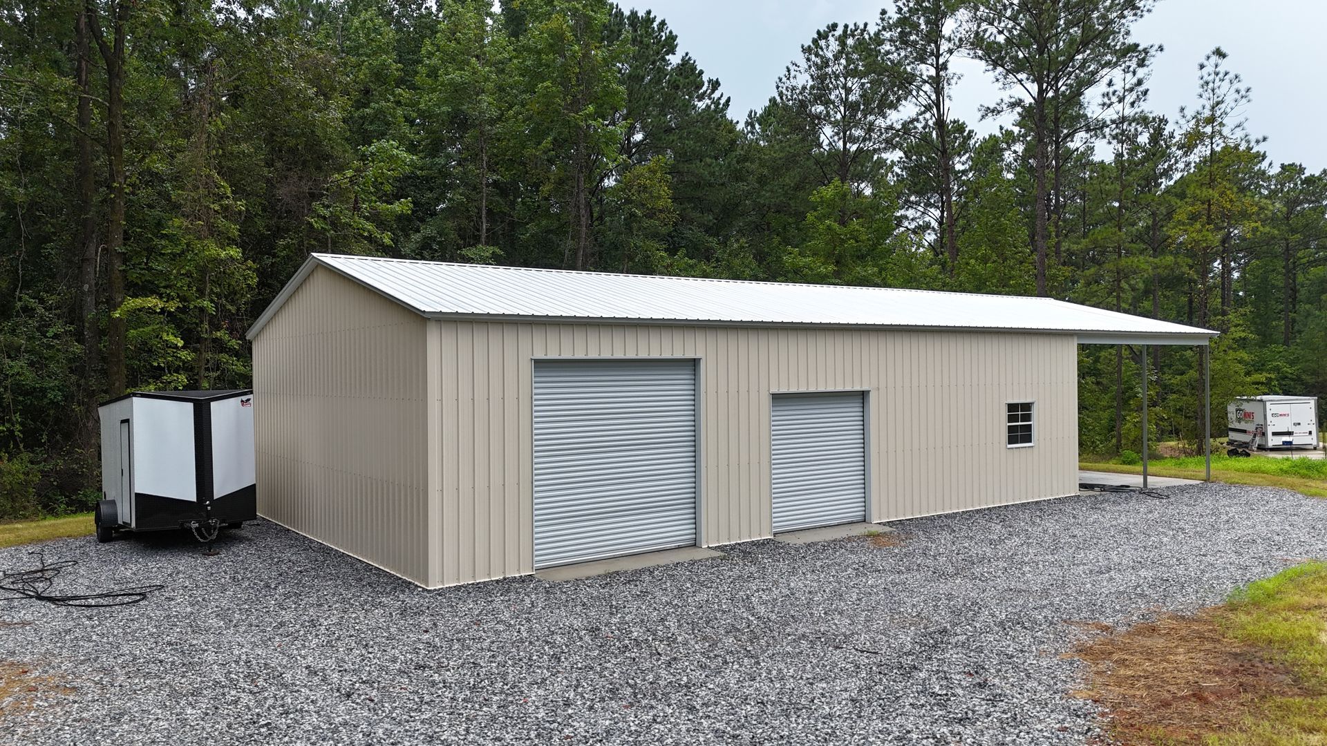Beige metal garage building with two roll-up doors on a gravel lot, surrounded by trees