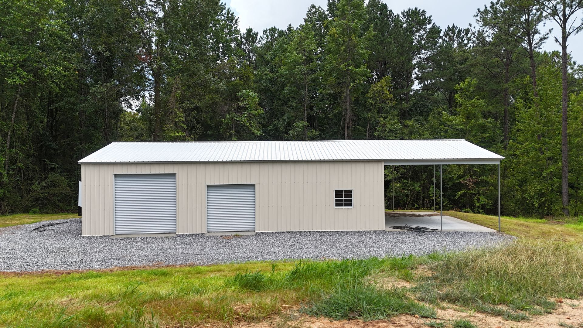 Small beige metal garage with two white roll-up doors and a covered side parking area, set by trees and gravel.