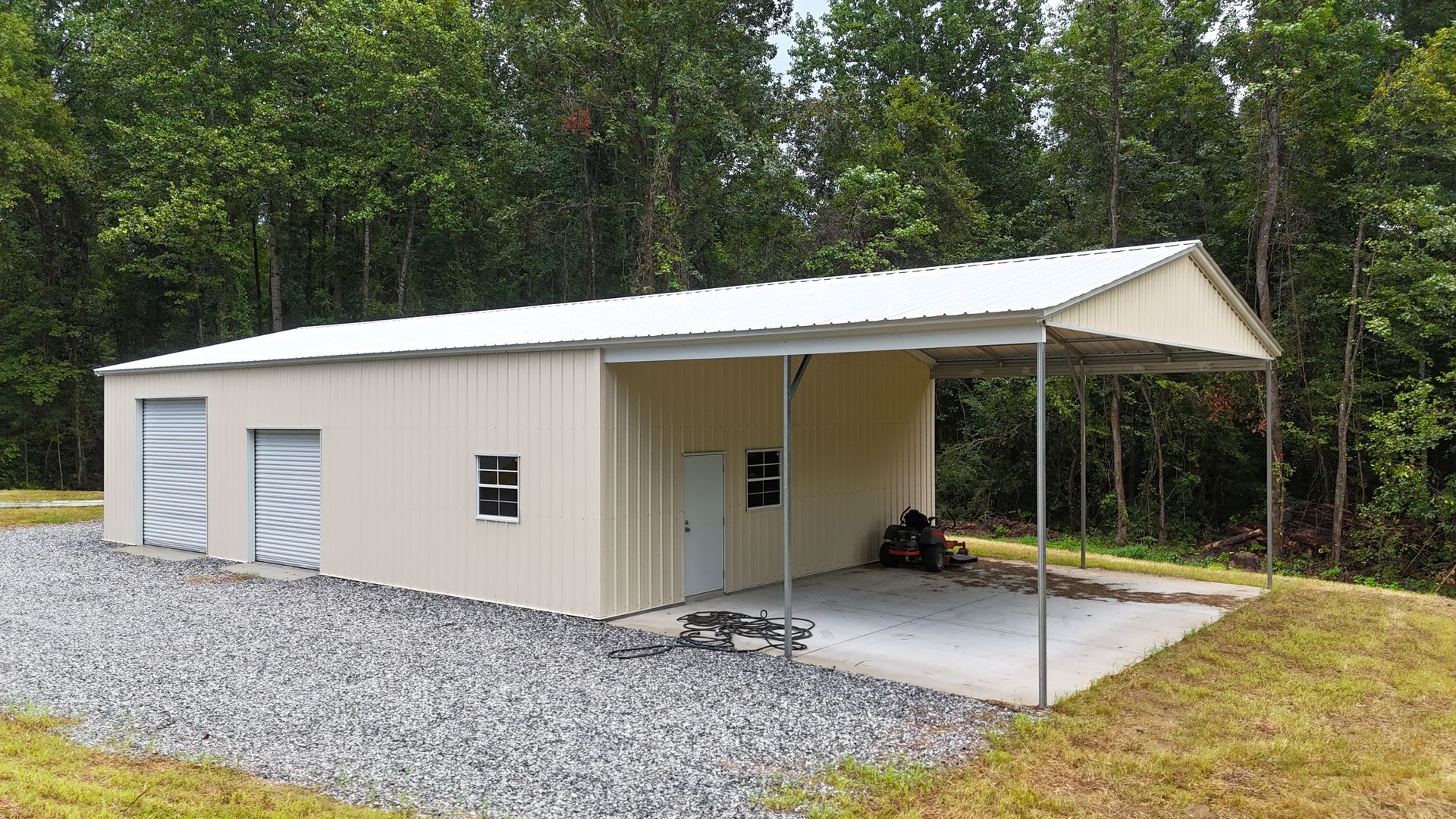 Tan metal garage with two roll-up doors and a covered carport beside a wooded yard