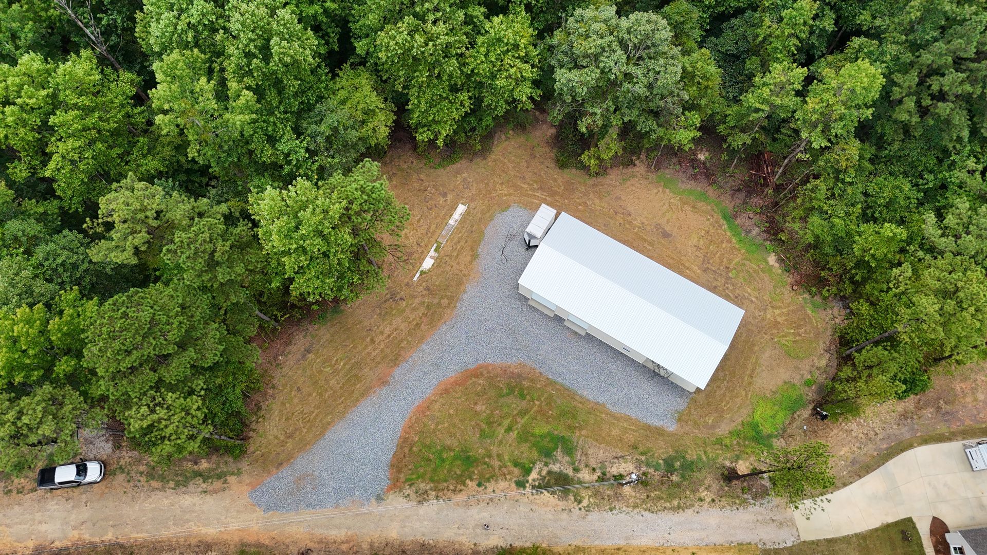 Aerial view of a white-roofed shed beside a gravel driveway at the edge of a wooded lot.