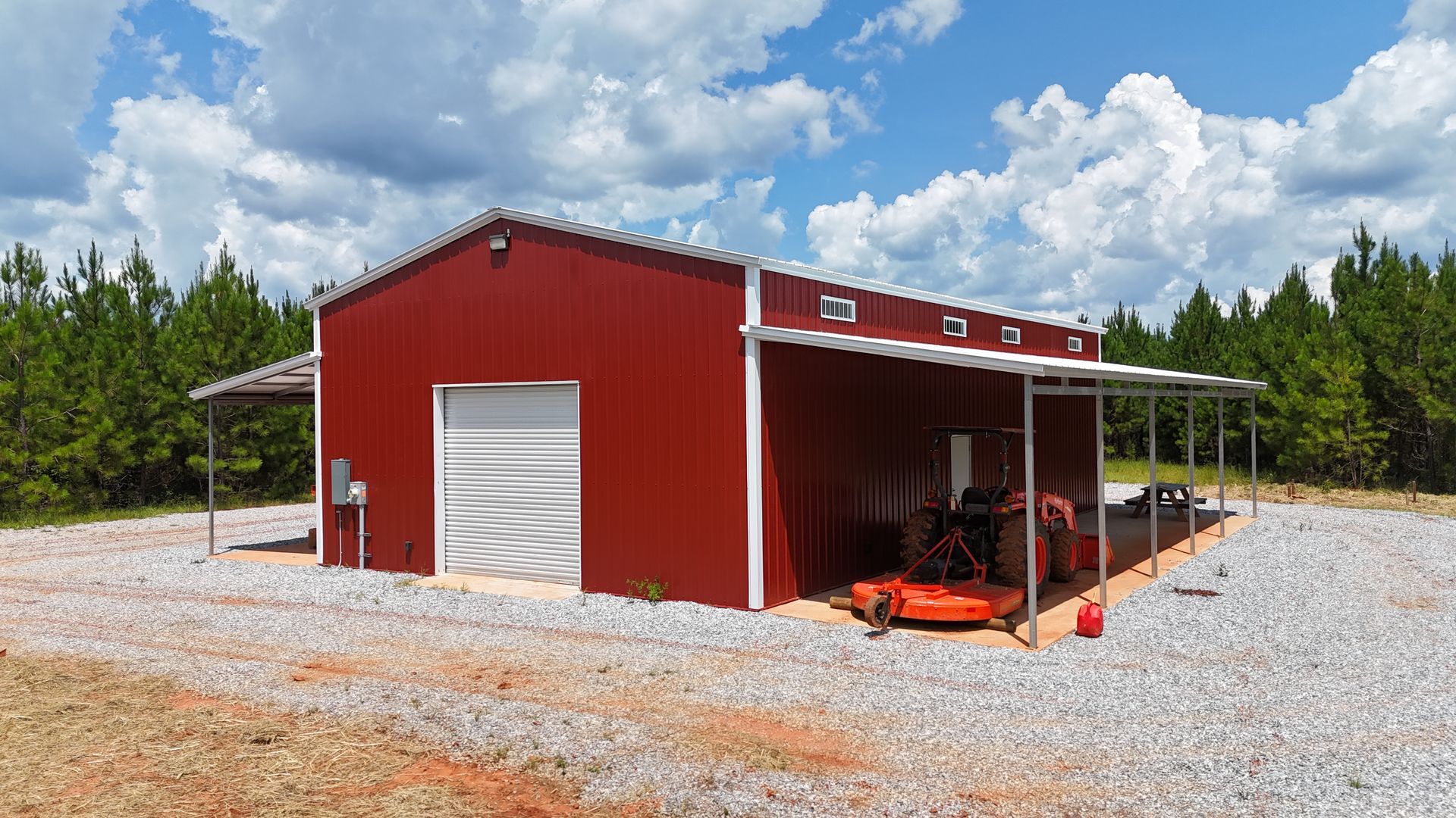 Red metal barn with open side bay on a gravel lot under a cloudy sky