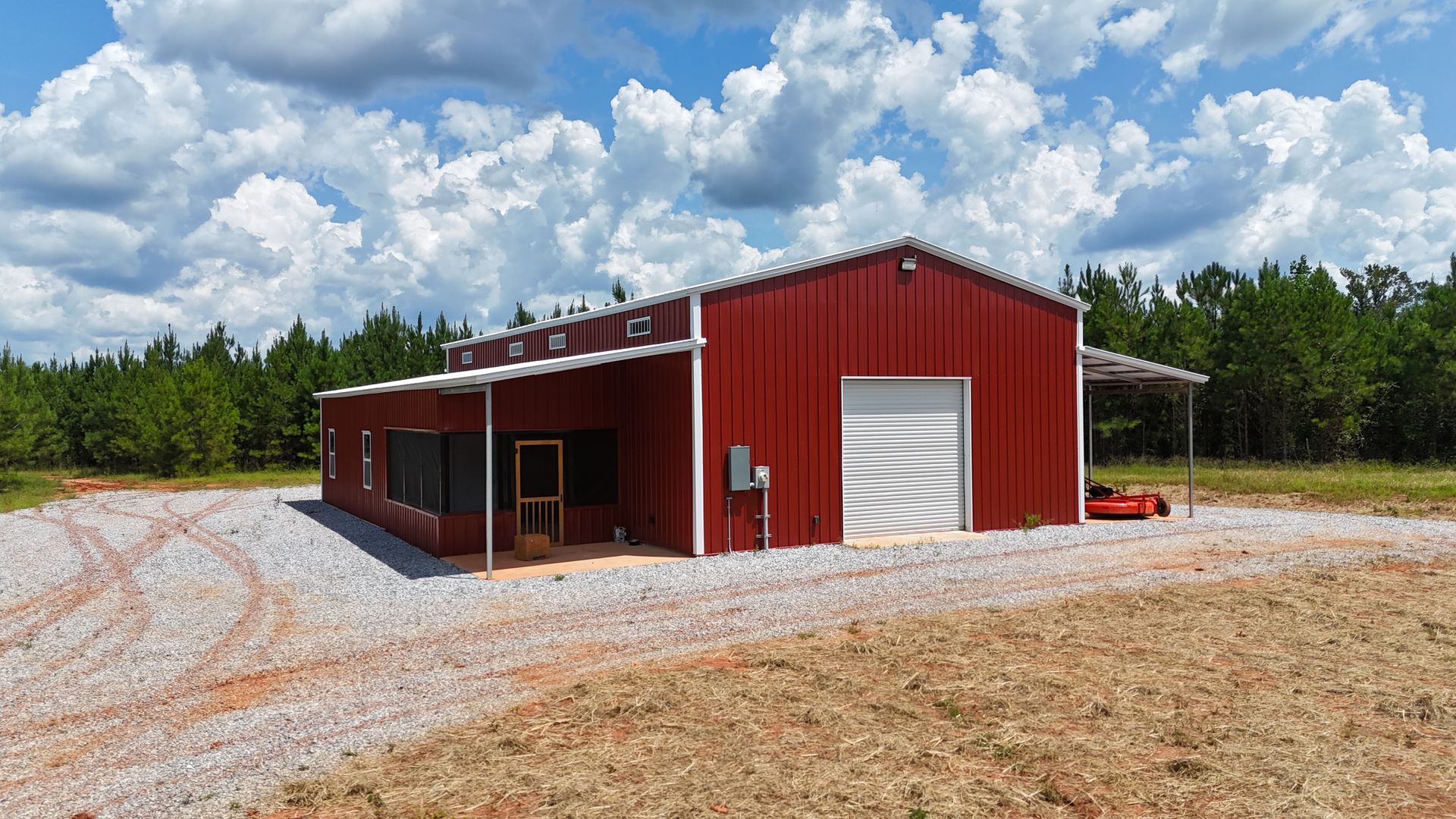Red metal workshop building with white garage door on gravel lot under a cloudy sky