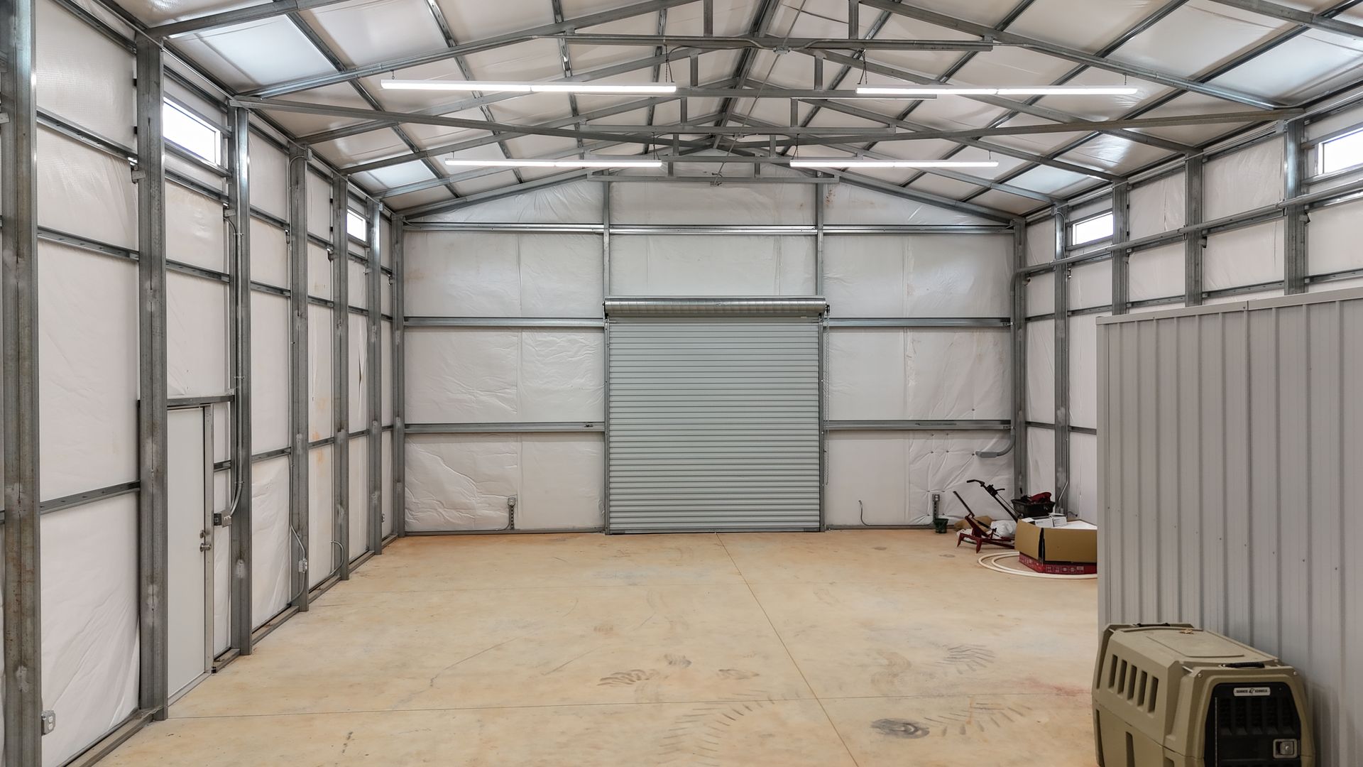 Empty metal storage shed with concrete floor, a roll-up door, and a few items stacked along the right wall.