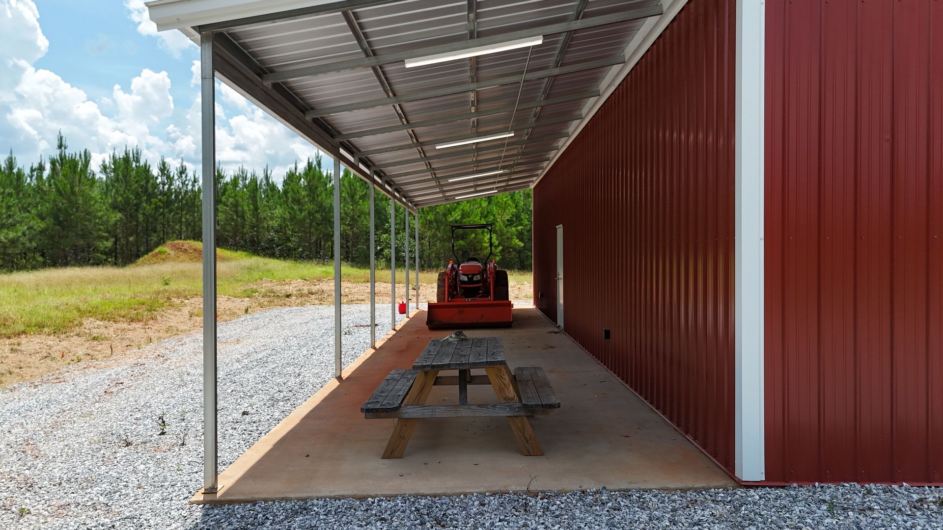Covered outdoor workspace beside a red storage container, with picnic tables on gravel under a metal roof.