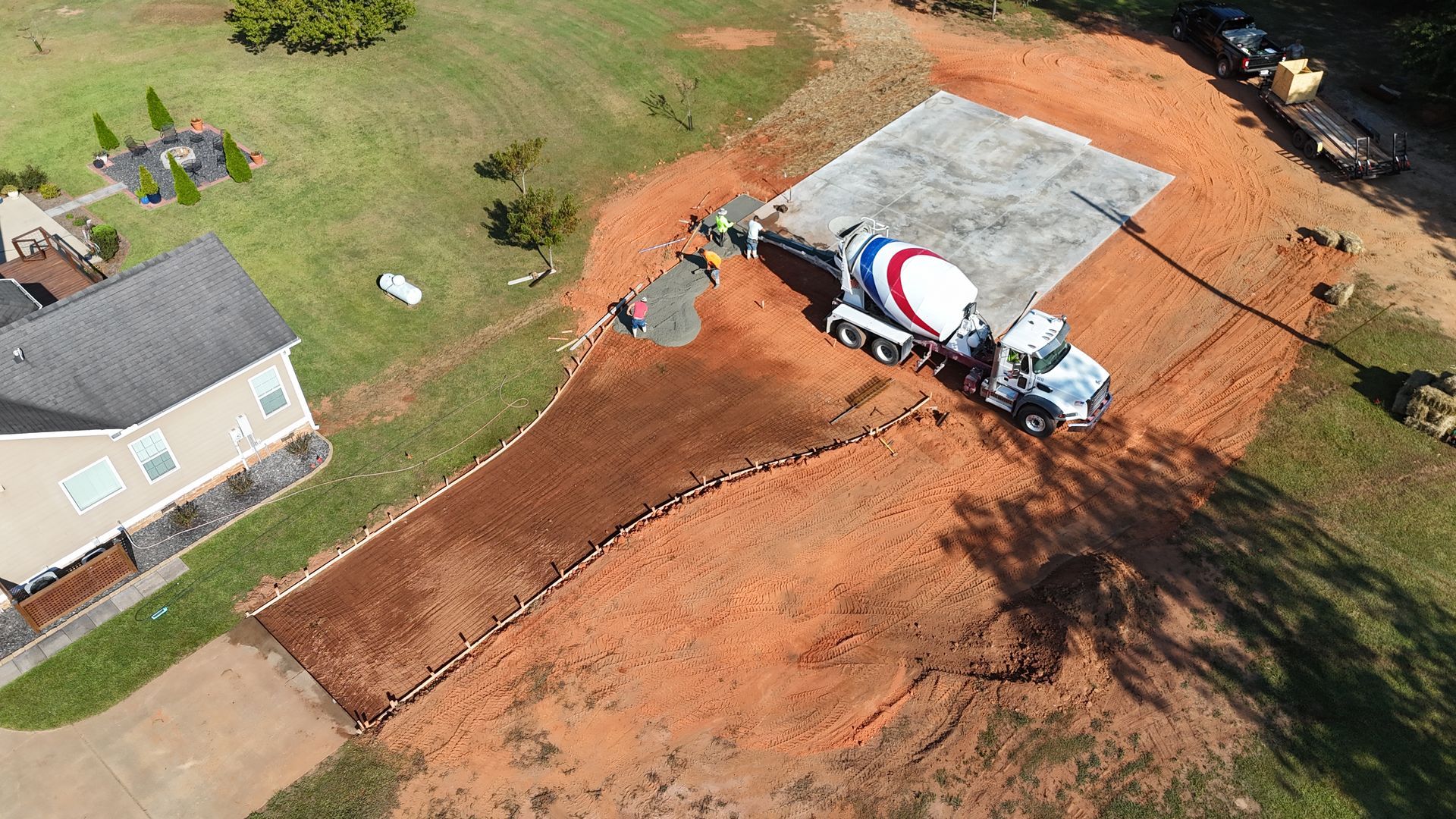 Aerial view of a concrete truck pouring a driveway beside a house, with fresh red dirt and a tarp-covered area.