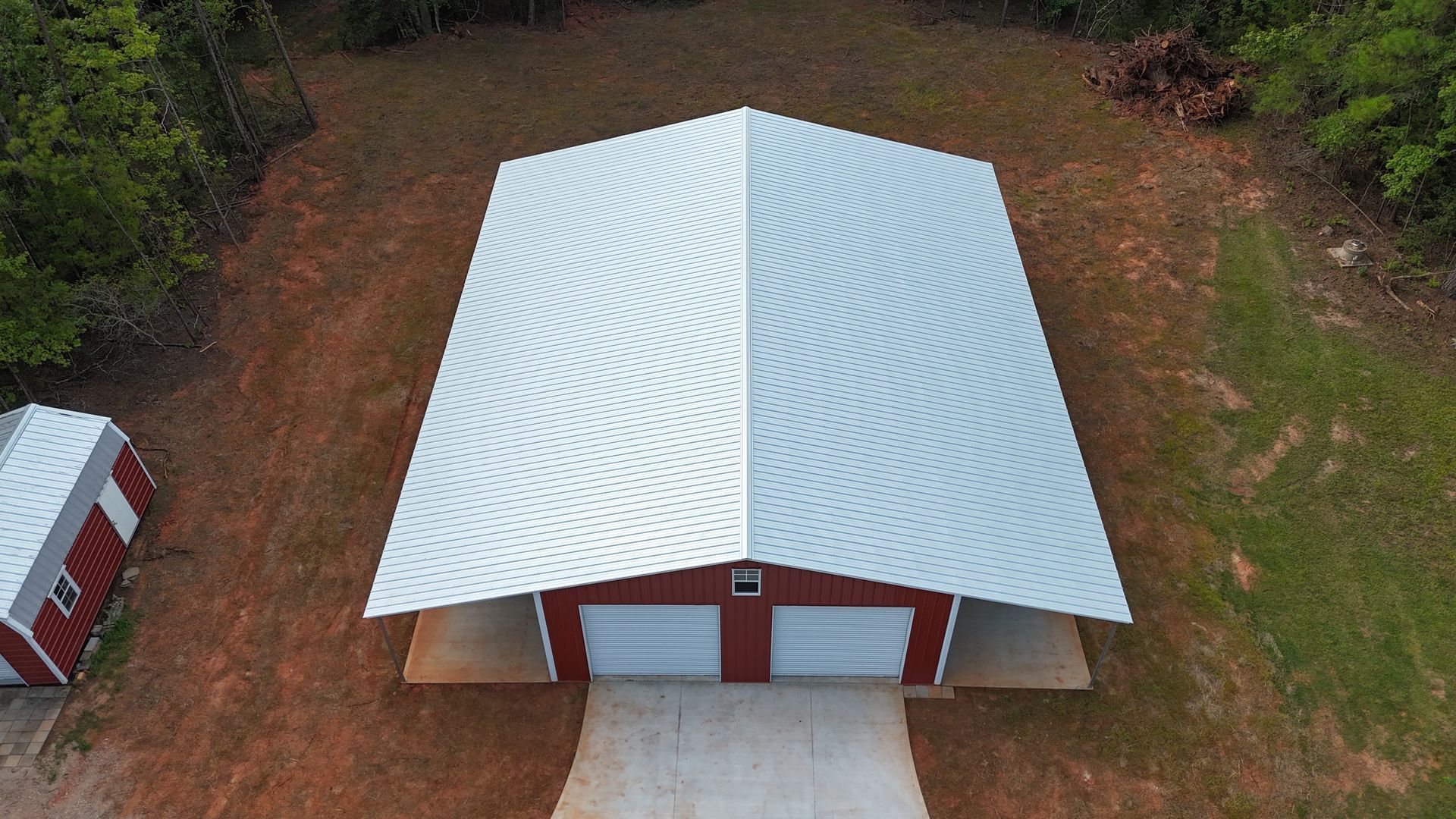 Aerial view of a red barn with a white metal roof and concrete driveway, surrounded by trees and dirt.