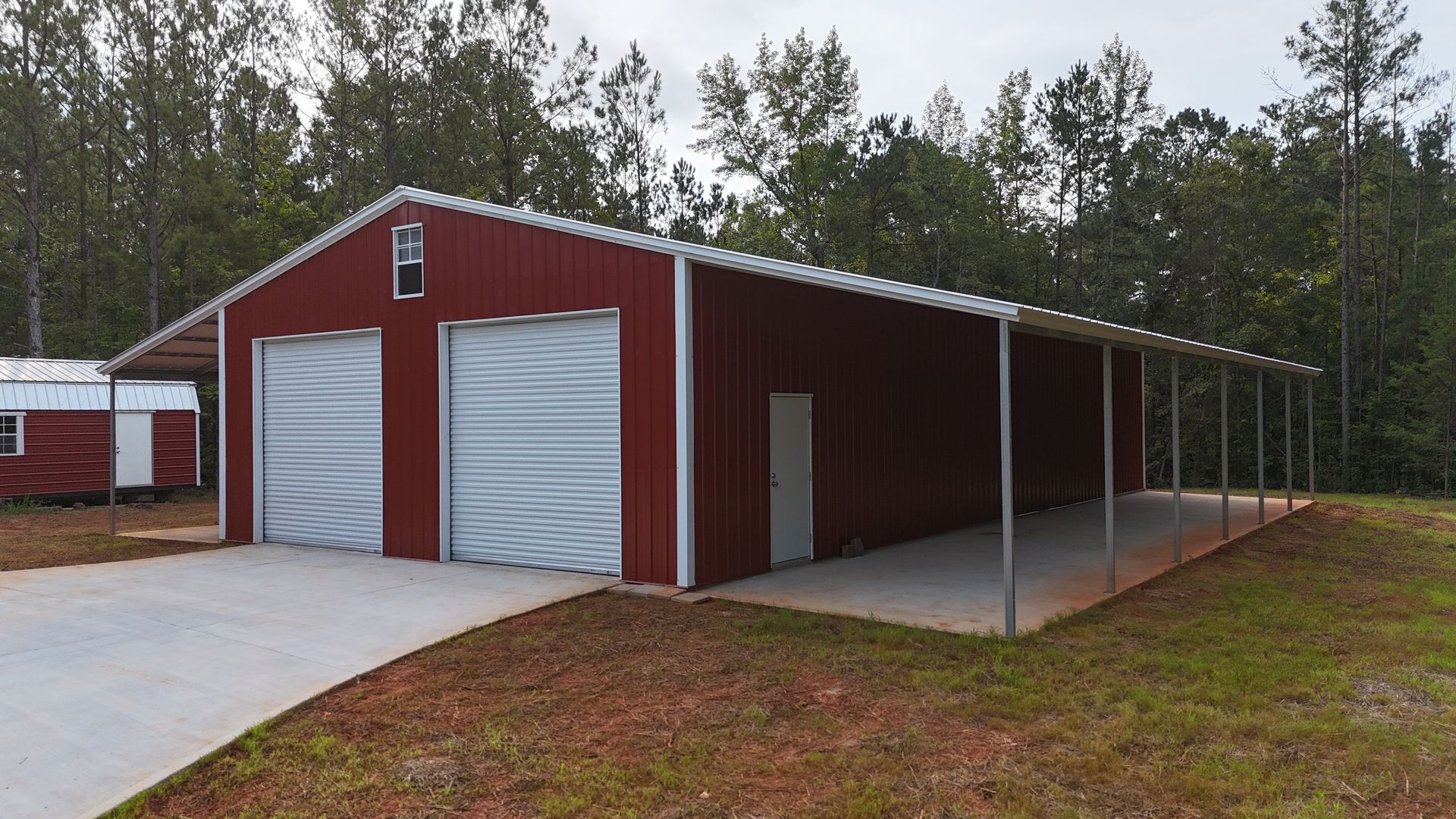 Red metal barn with white roll-up doors and a covered side awning beside a gravel drive and trees