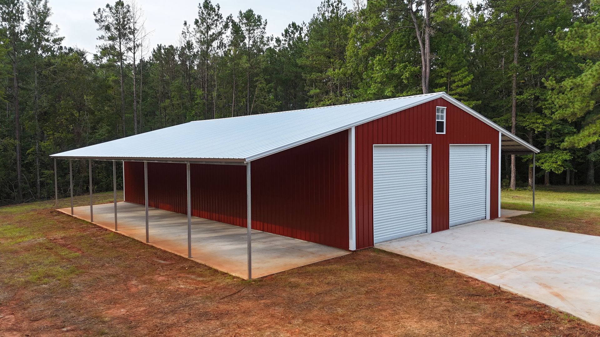 Red metal barn with white roof, two garage doors, and covered porch on a wooded lot.