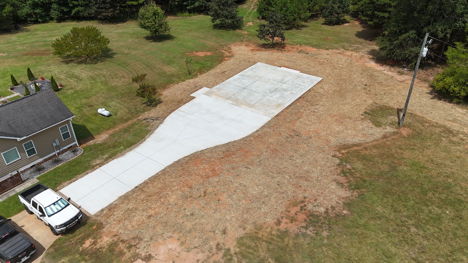Aerial view of a driveway with a white concrete slab beside a house and parked truck, surrounded by trees and grass