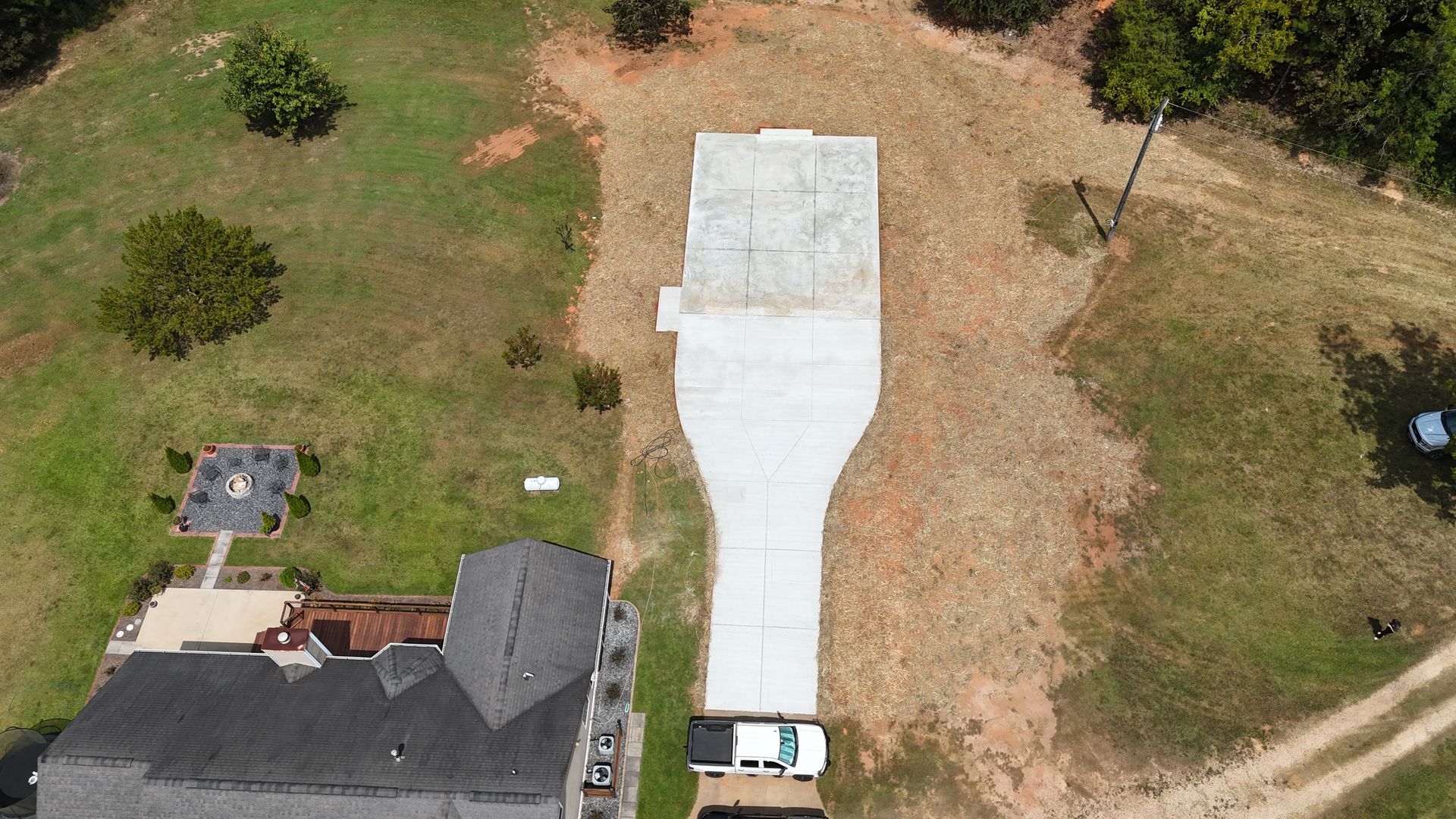 Aerial view of a house with a long white driveway and surrounding grassy yard, with a car at the bottom
