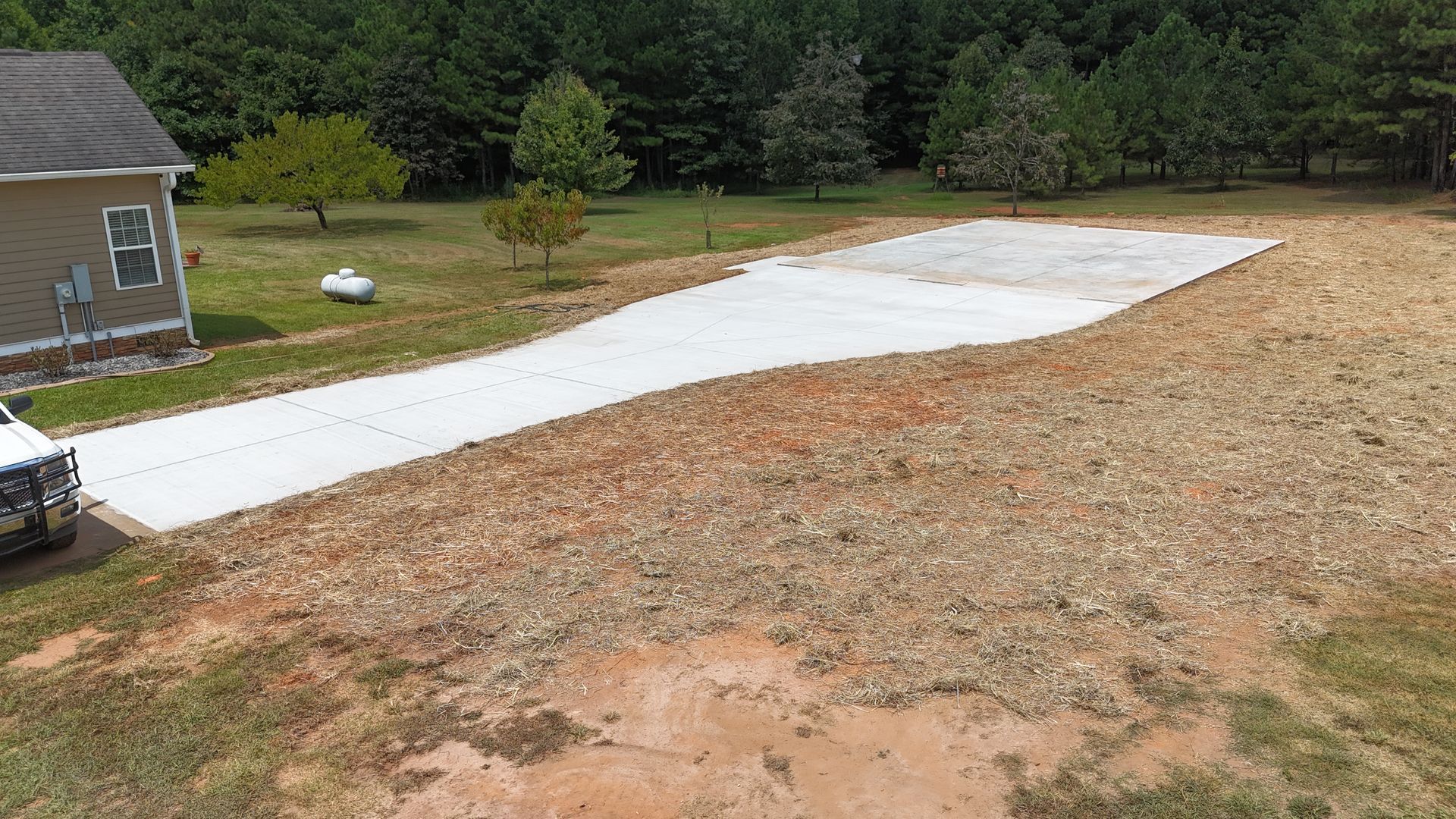 Freshly poured white concrete driveway beside a house, stretching across a dirt lot toward the woods.