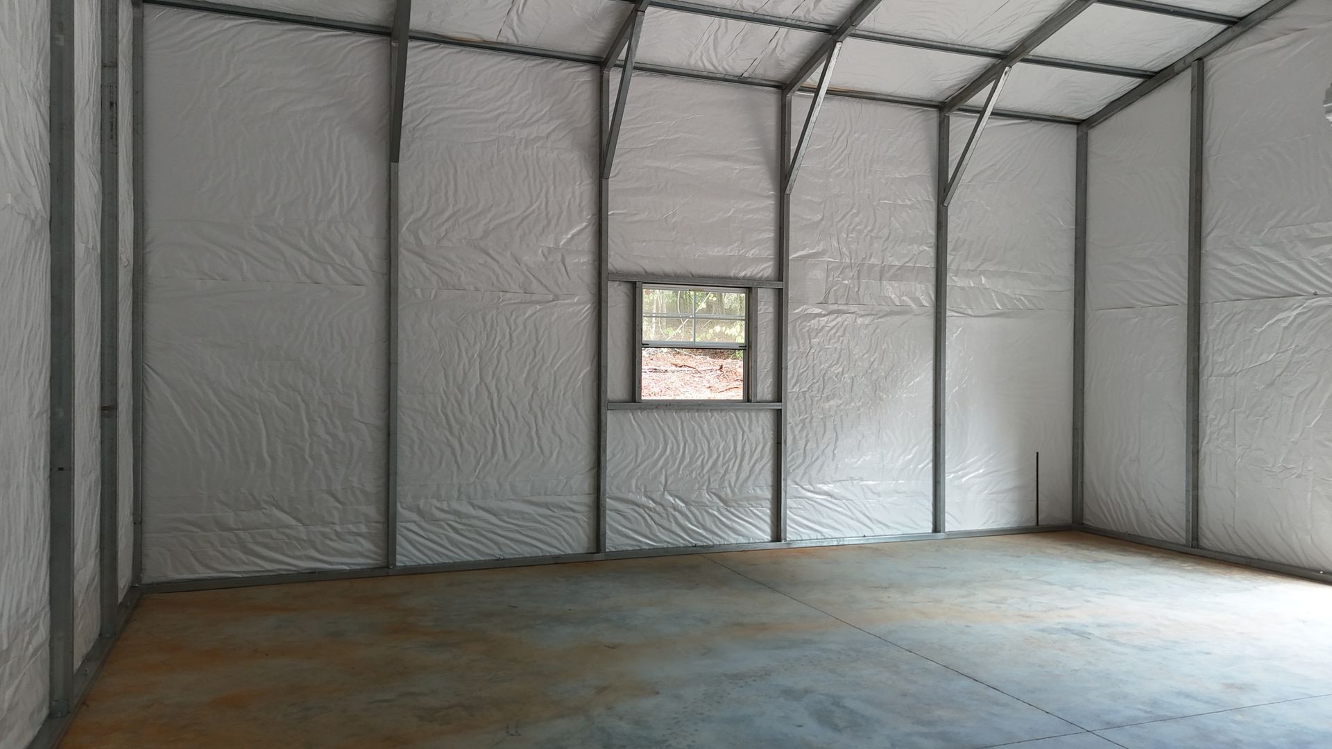 Empty insulated room with metal framing, white sheeting, concrete floor, and a small window.