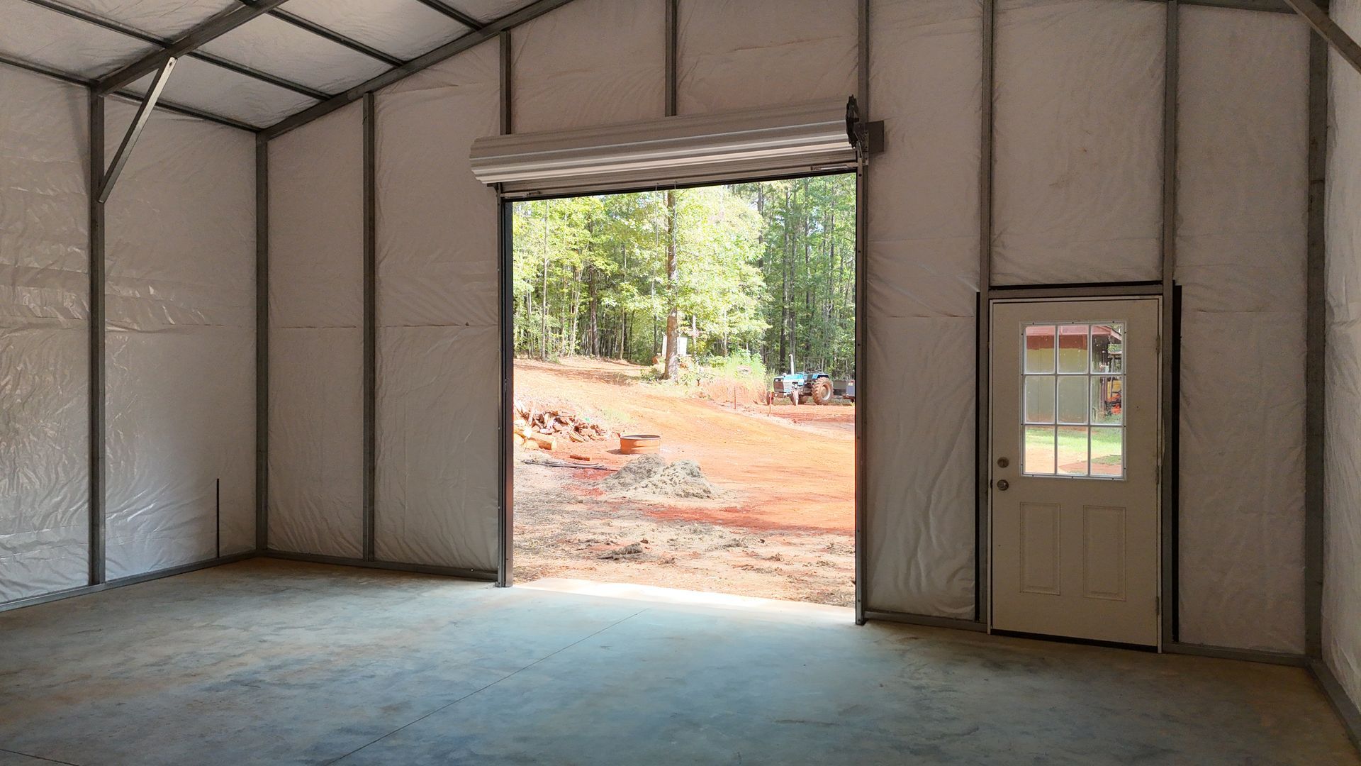 Empty metal building interior with open roll-up door, concrete floor, and a small windowed door on the right.