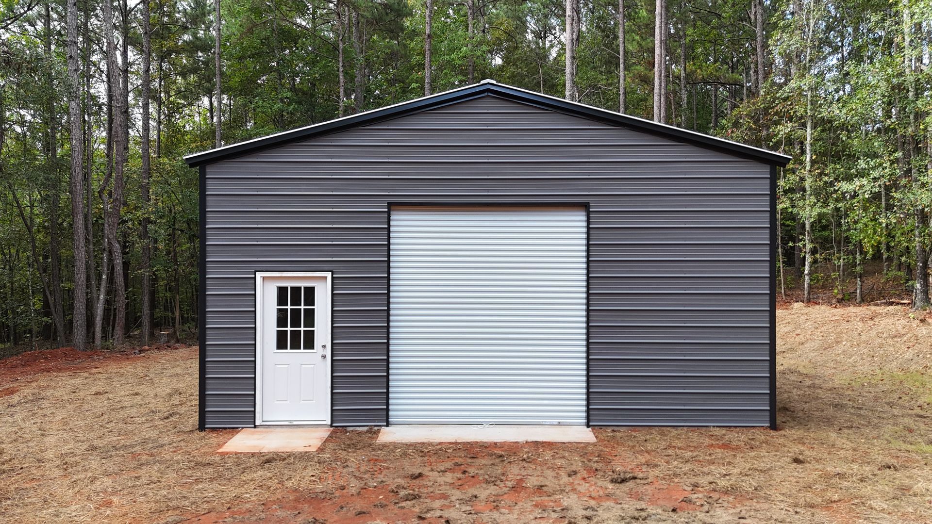 Gray metal storage building with a white roll-up door and side entry door in a wooded lot