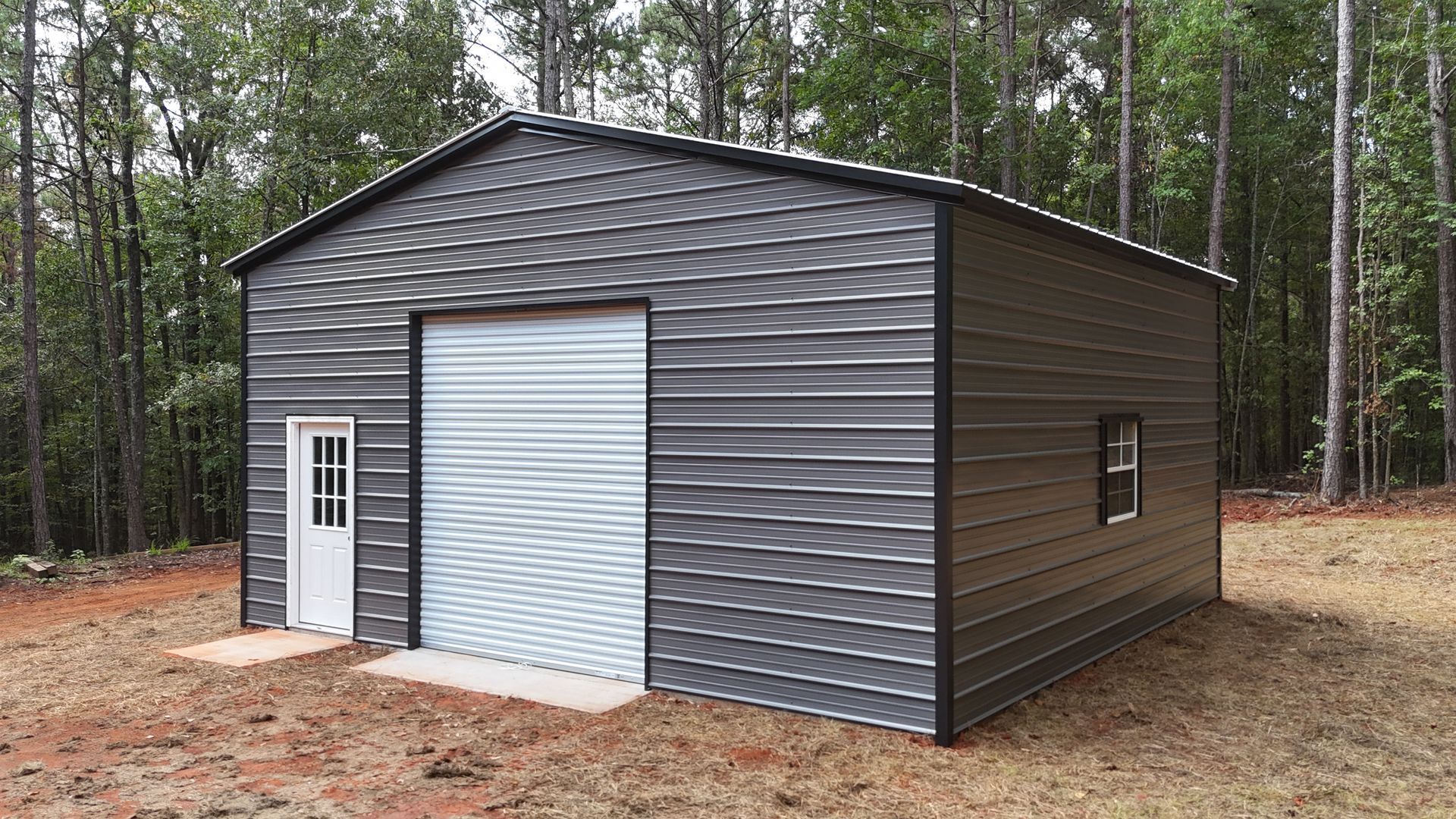Dark gray metal storage building with a roll-up door and side entry door in a wooded clearing