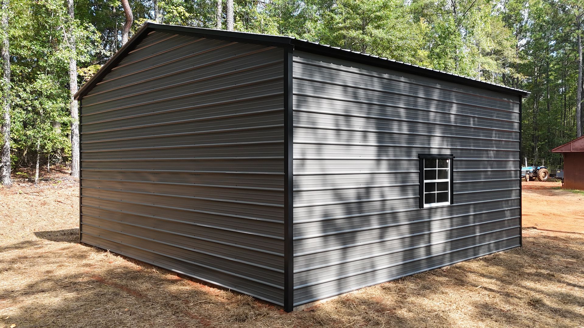 Dark gray metal shed with a small window, set on a wooded lot with trees and leaf-covered ground.