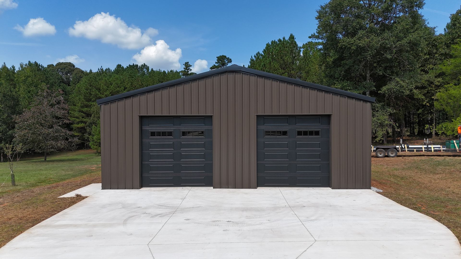 Brown metal garage with two black doors on a concrete pad, surrounded by trees and grass under a blue sky
