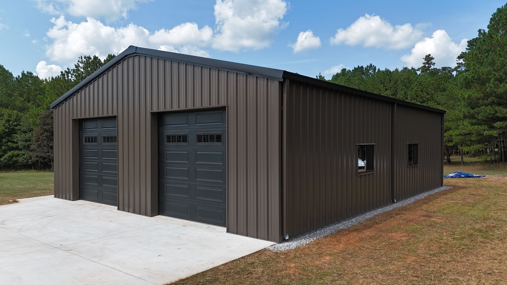 Dark metal workshop with two garage doors on a concrete slab beside trees and grass under a cloudy sky