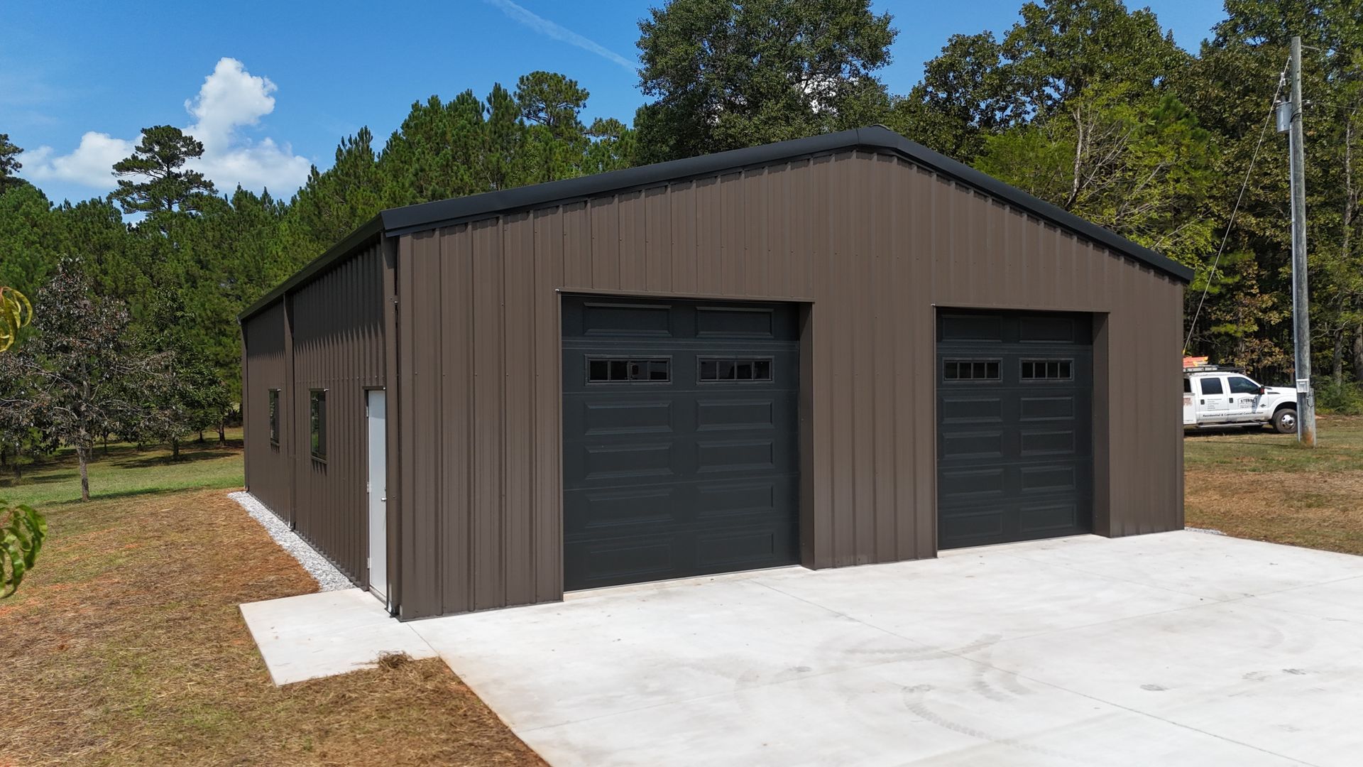 Brown metal garage with two dark doors on a concrete driveway, surrounded by trees under a clear sky