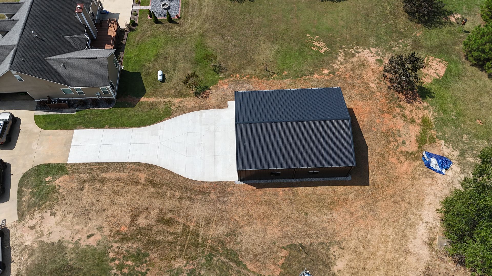 Aerial view of a small dark-roofed shed beside a white concrete driveway on a grassy lot.