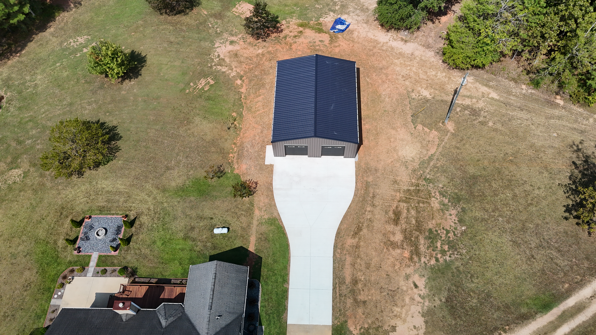 Aerial view of a small blue-roofed shed with a white driveway in a grassy yard, beside a house and trees