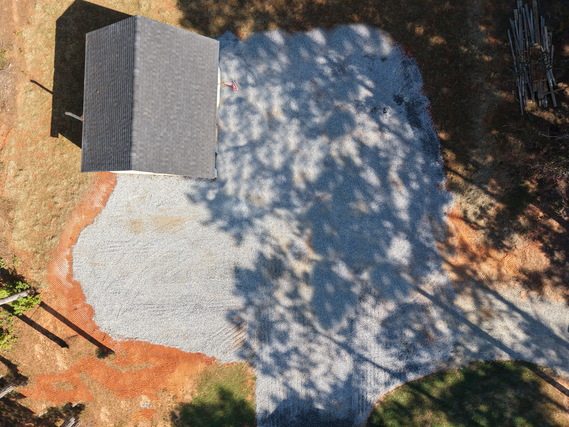 Overhead view of a gravel driveway next to a building with a dark gray roof, and trees casting long shadows.