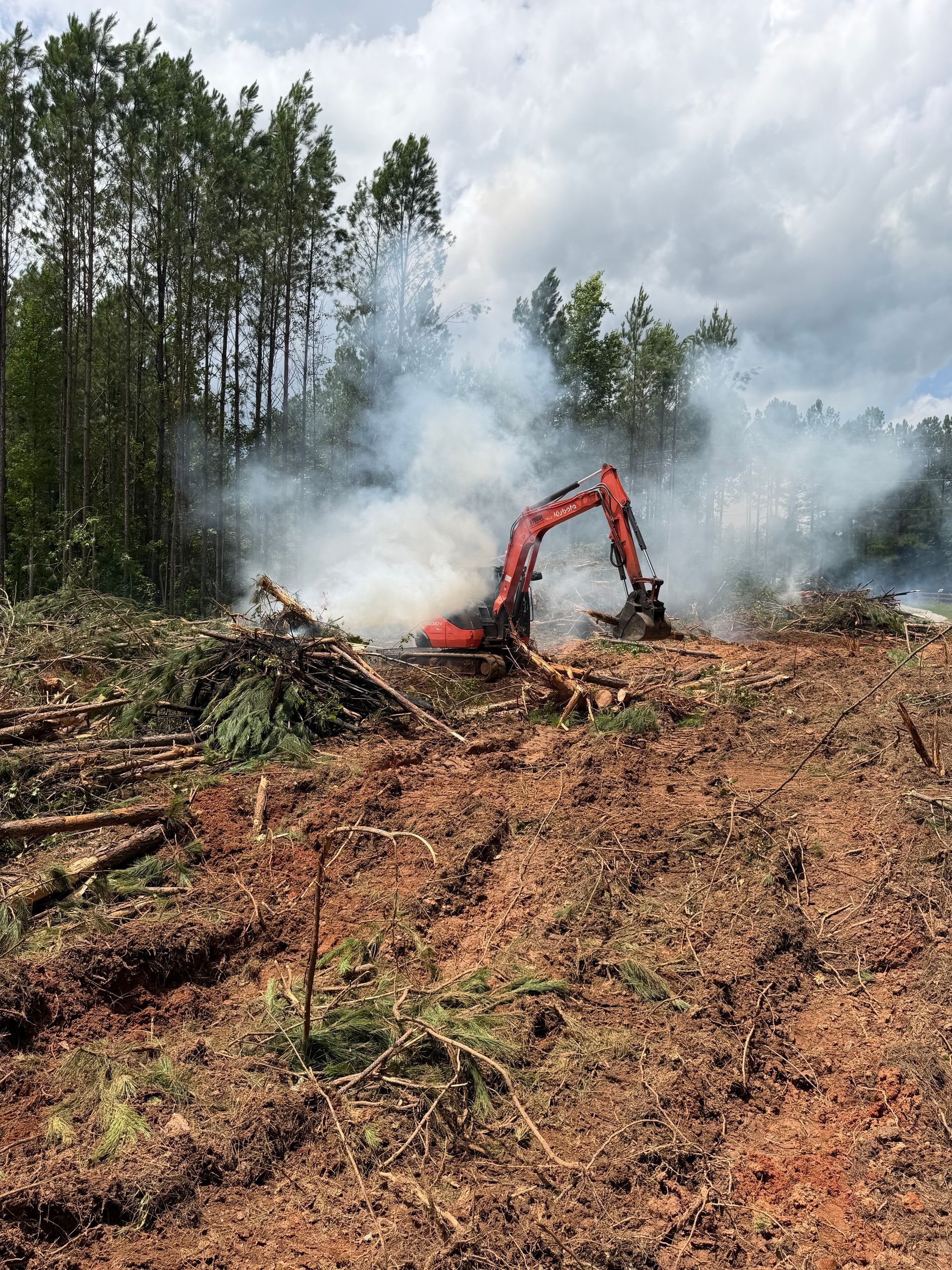 Red excavator clearing a smoky forest area with felled trees and muddy ground